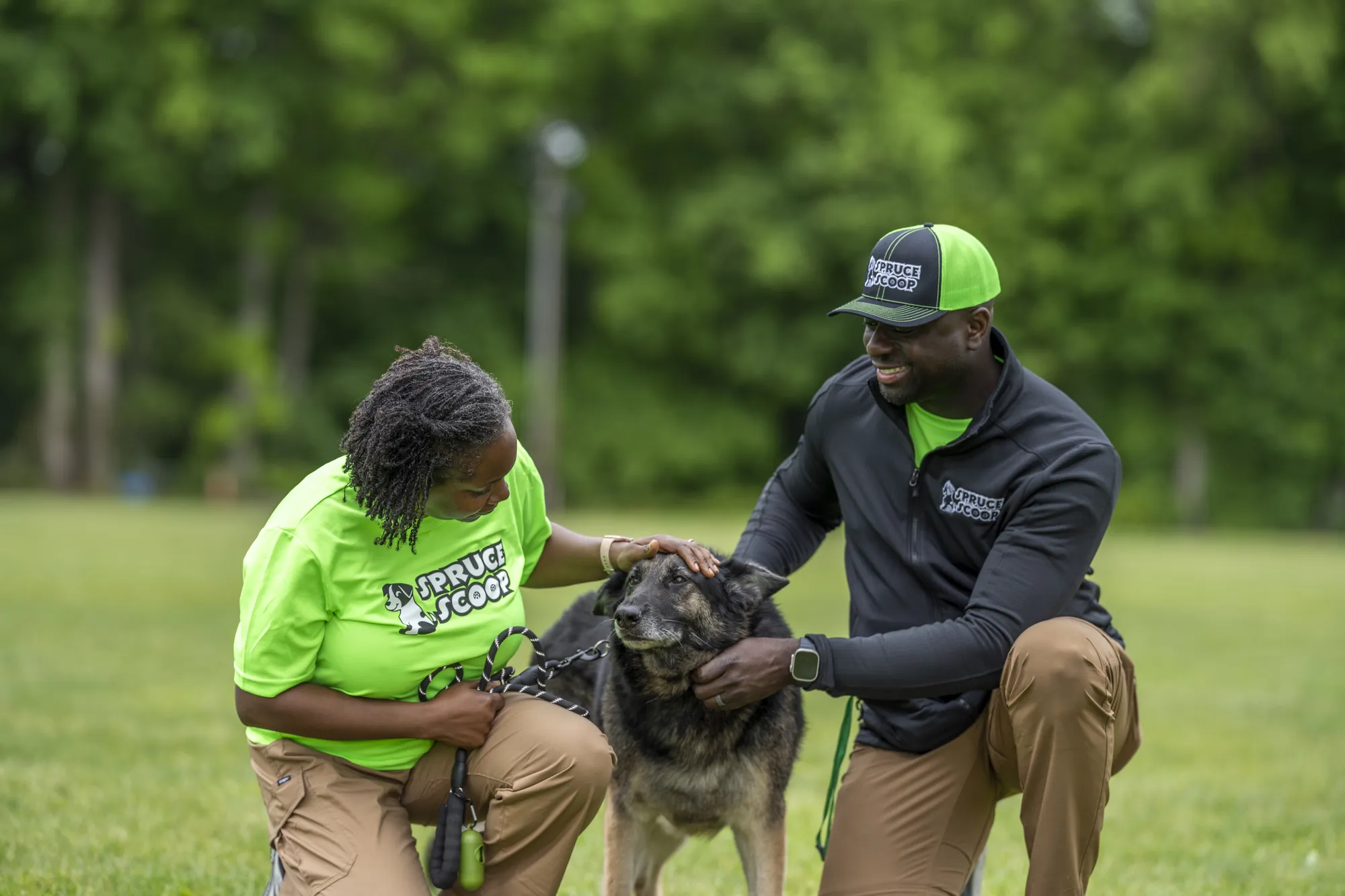 Two Spruce Scoop team members kneeling to pet a dark dog on a grassy field