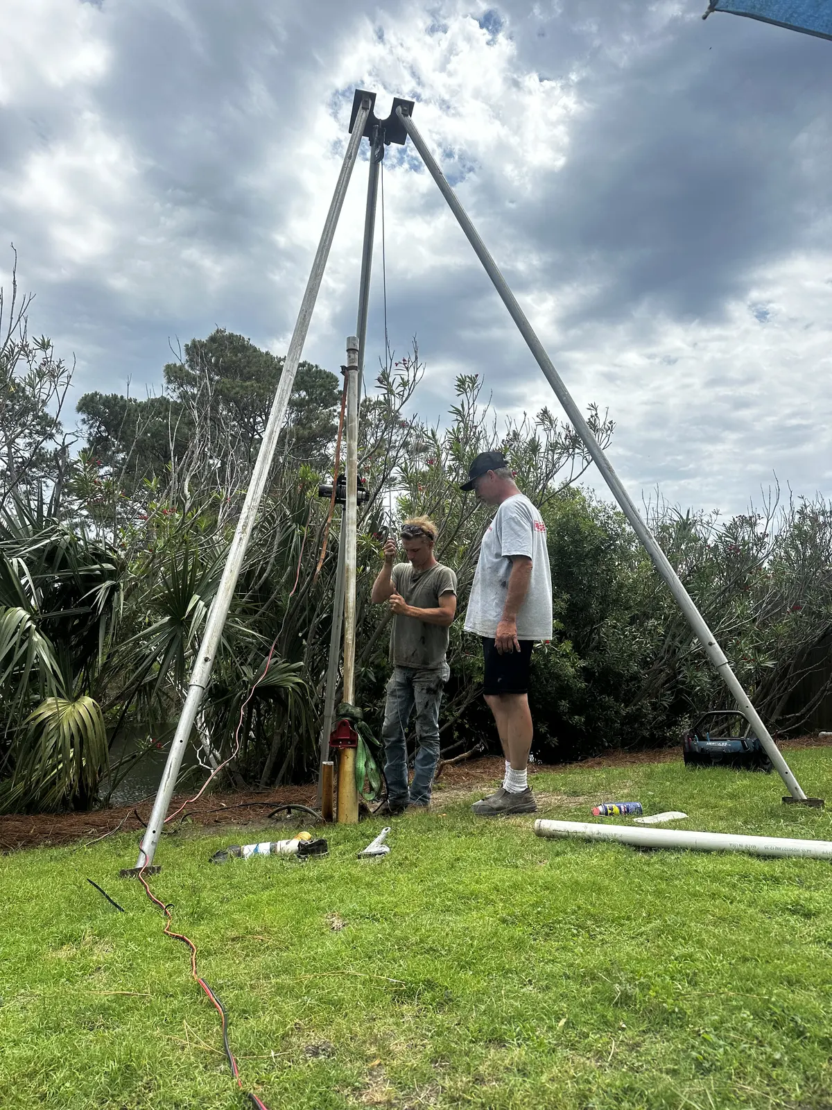 Two crew members operating a tripod exploratory drilling rig in a grassy field