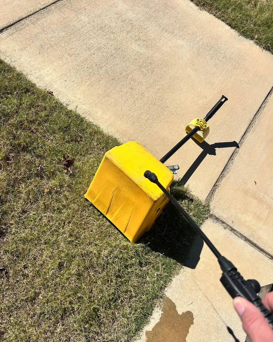 A yellow scoop and a rake on the grass beside a sidewalk, with a hand holding a sprayer wand