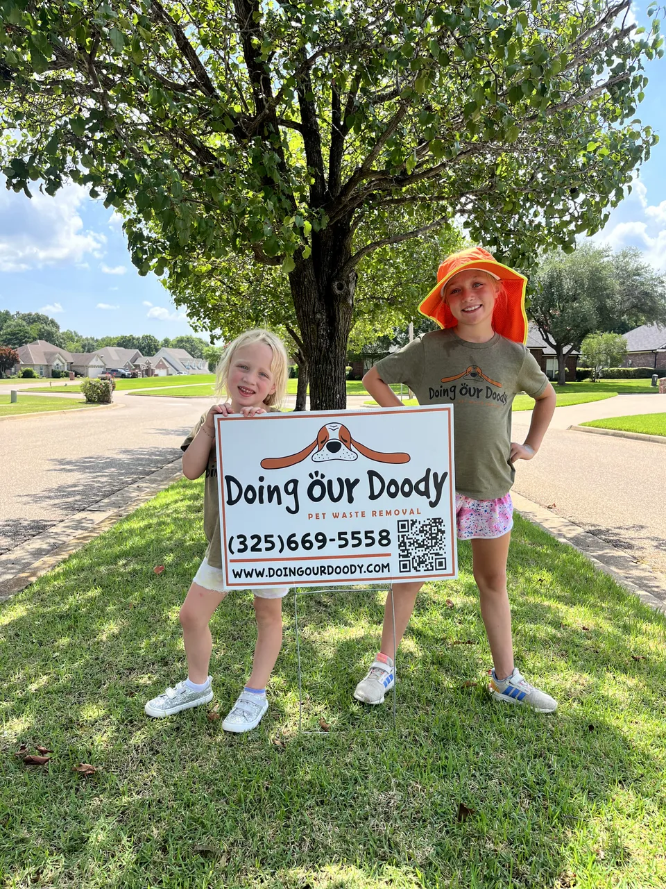 Two young girls smiling in front of a Doing Our Doody pet waste removal sign