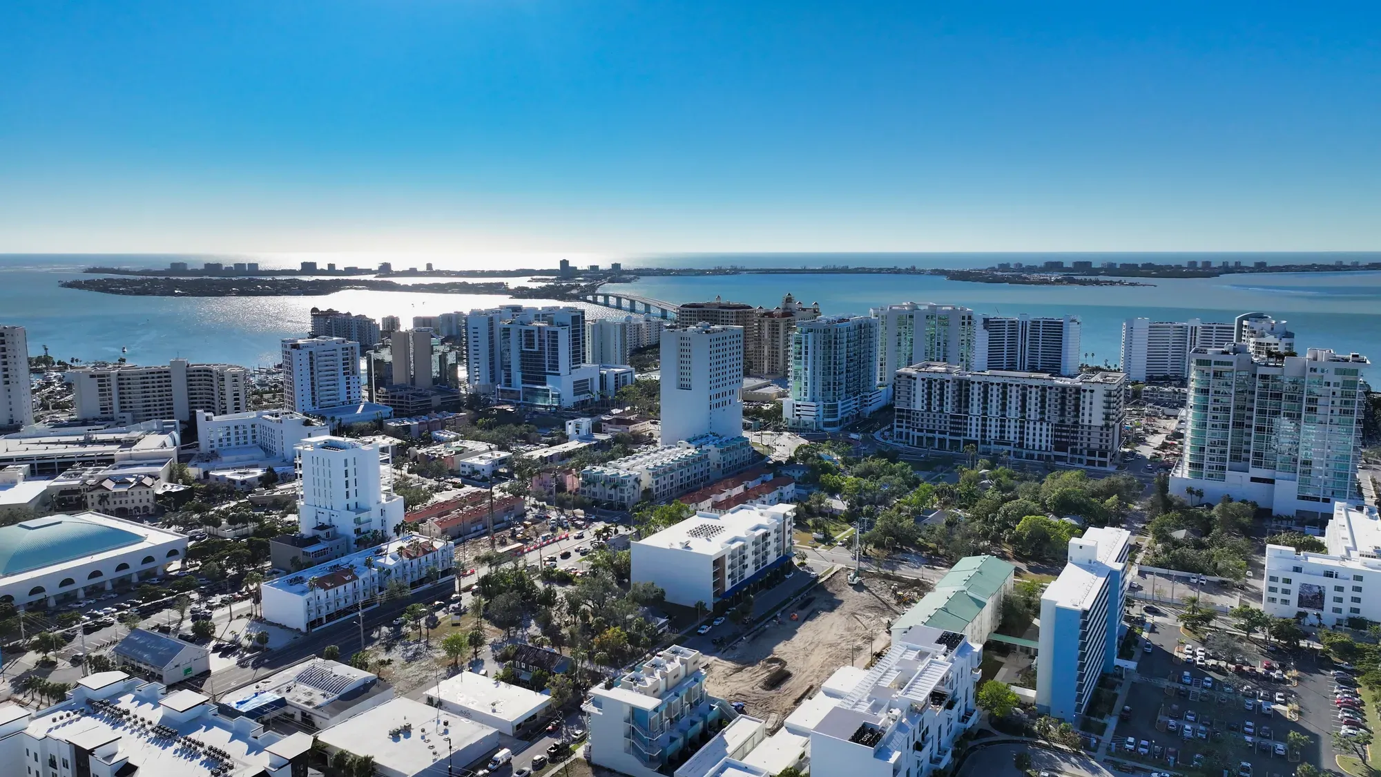 This aerial image showcases a modern waterfront city with high-rise buildings and a scenic body of water in the background. The bright blue sky and reflection on the water enhance the vibrant, urban atmosphere.