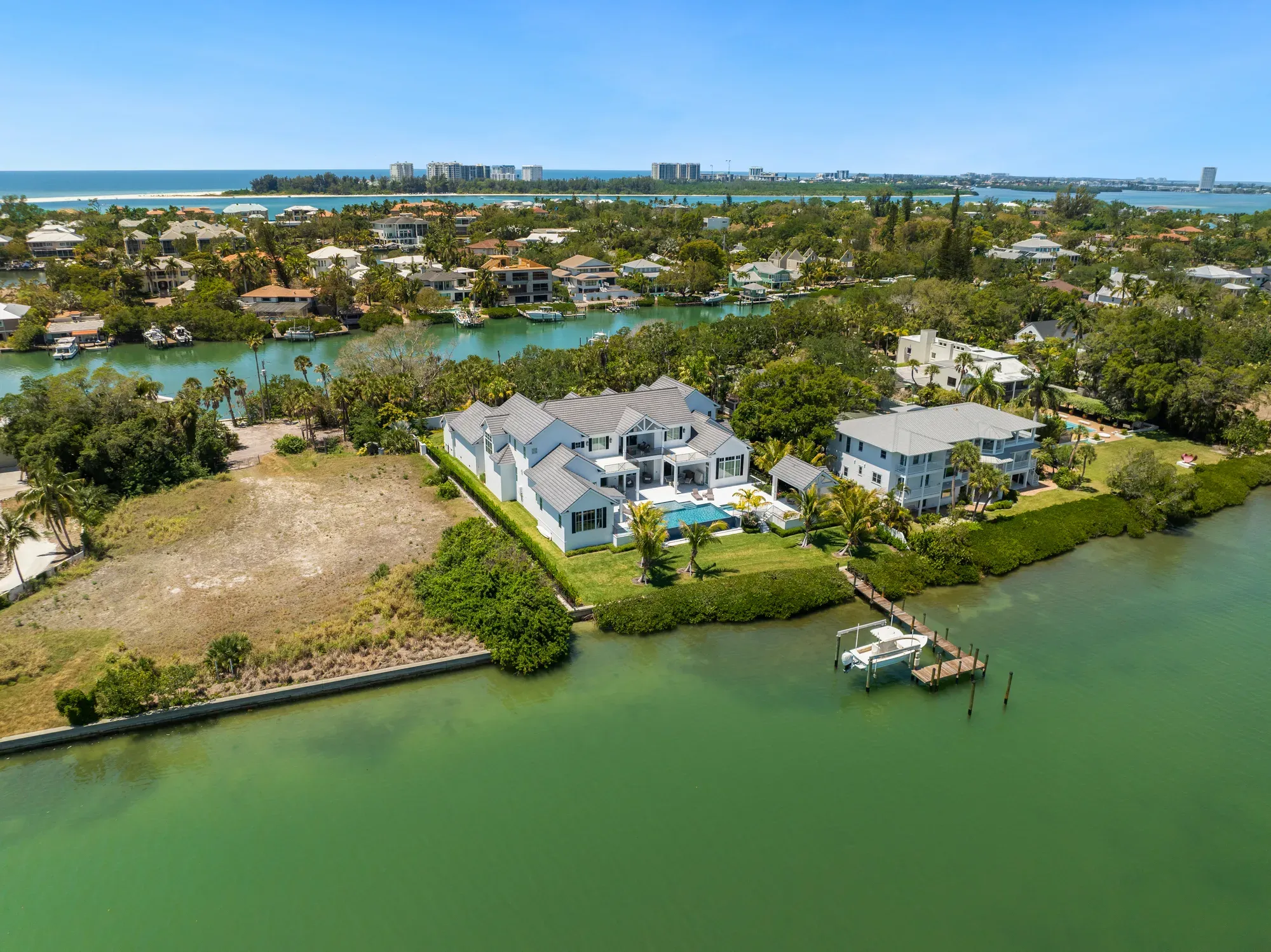 This aerial image captures a residential area with waterfront homes, lush greenery, and clear blue waters. Notable features include a spacious house with a pool and a dock, set against a backdrop of other homes and a distant view of the coastline.