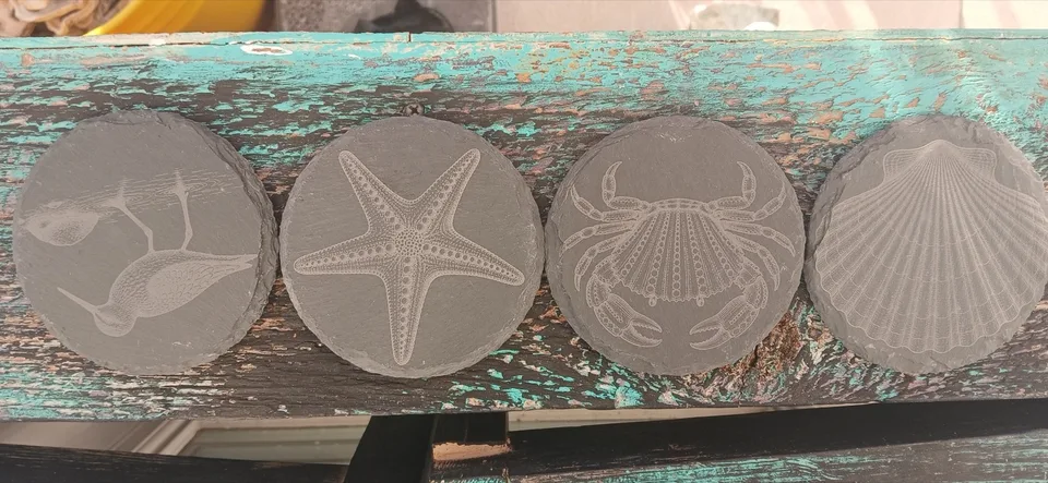 Four round slate coasters engraved with a shorebird, starfish, crab and scallop shell on a turquoise bench