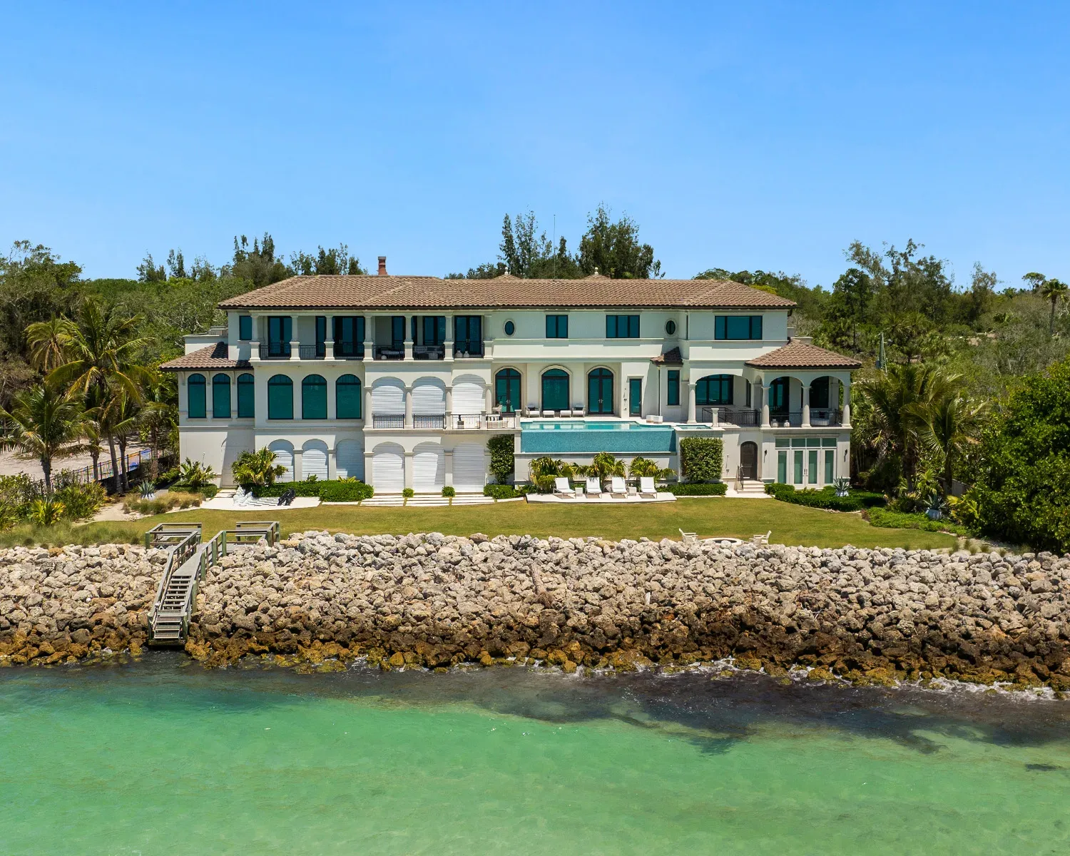 This image showcases a luxurious waterfront villa featuring a modern architectural design with a light color palette. Surrounded by palm trees, the property boasts a landscaped yard and direct access to the water, providing a serene coastal view.