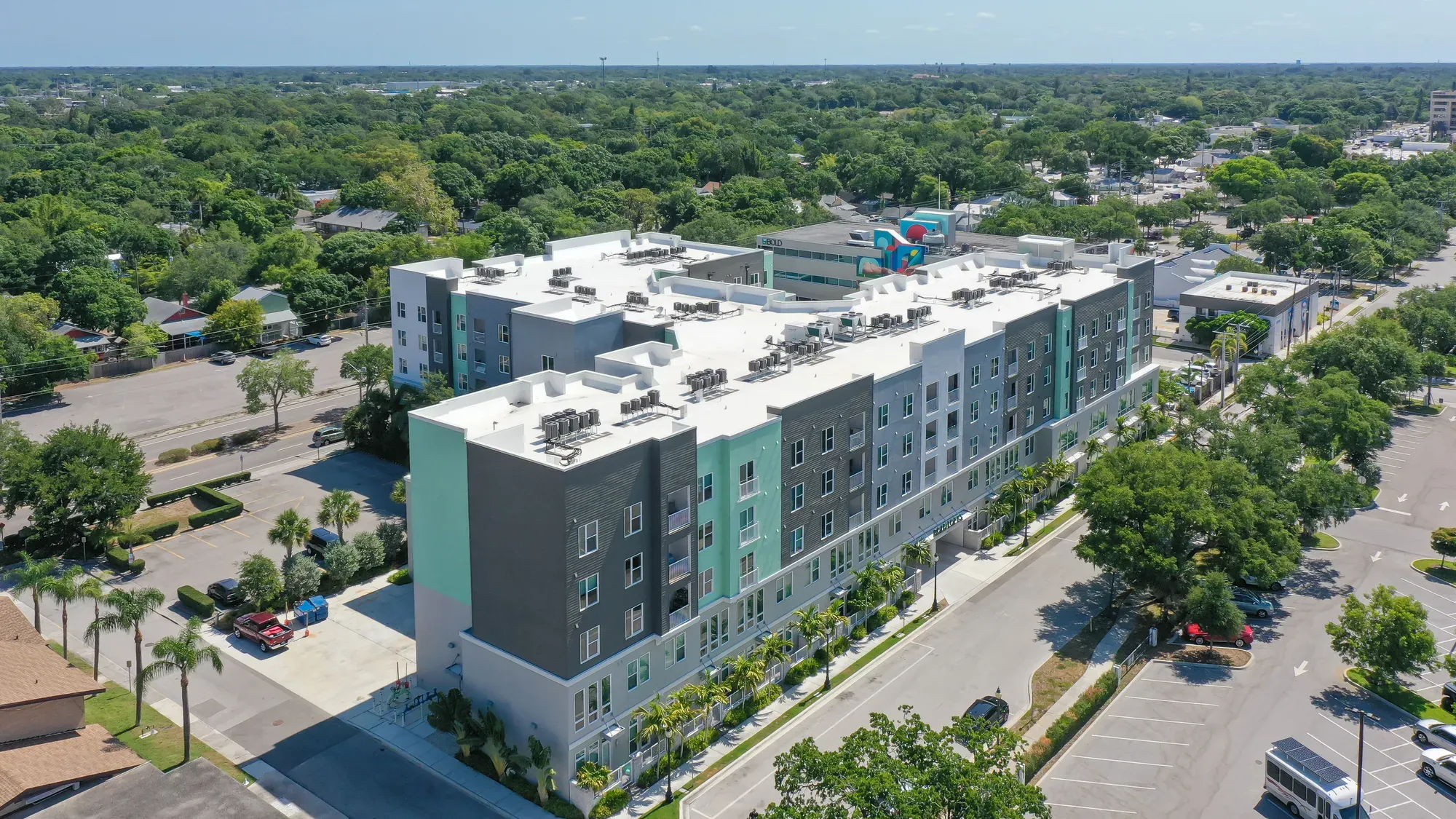 An aerial view of a modern apartment complex featuring multiple stories with a mix of colorful facades. Surrounding greenery and nearby roadways are visible, highlighting the residential area.
