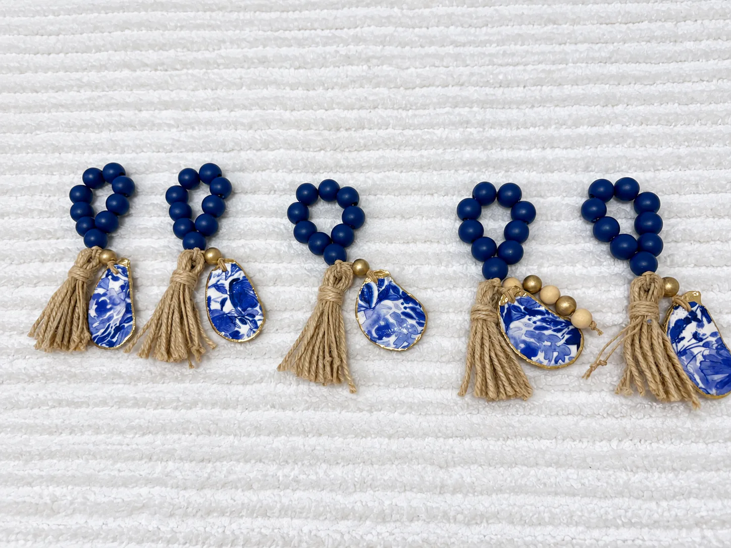 Five navy bead napkin rings with jute tassels and gold-edged oyster shell charms in a blue and white floral pattern