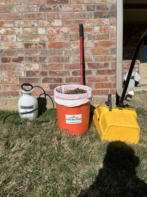 Red Doing Our Doody bucket, sprayer, rake, and scooper on grass against a brick wall
