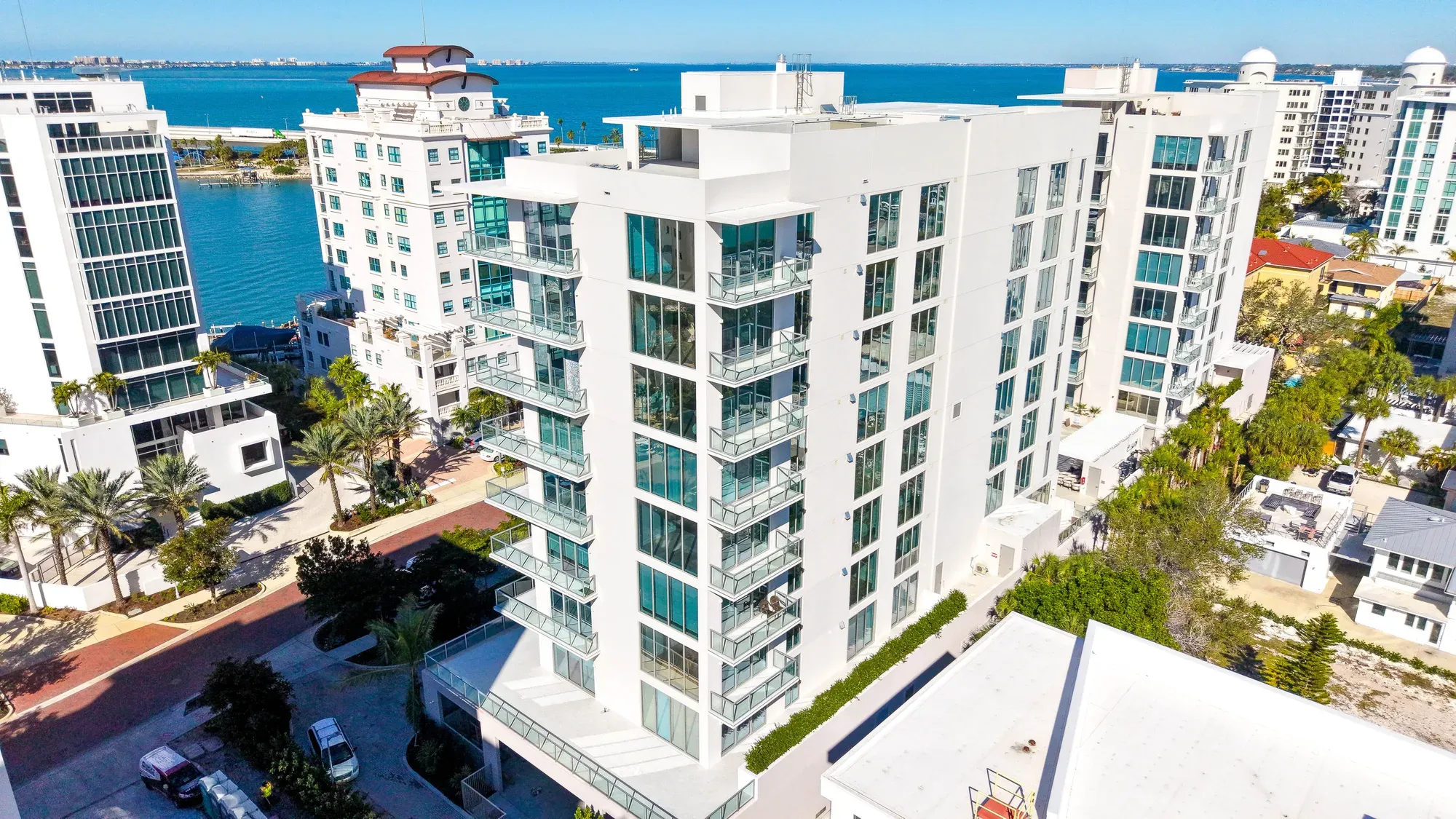 An aerial view of a contemporary multi-story apartment building with large glass balconies, situated near a serene waterfront. Palm trees line the pathways, and the backdrop features a clear blue sky and distant buildings along the shoreline.