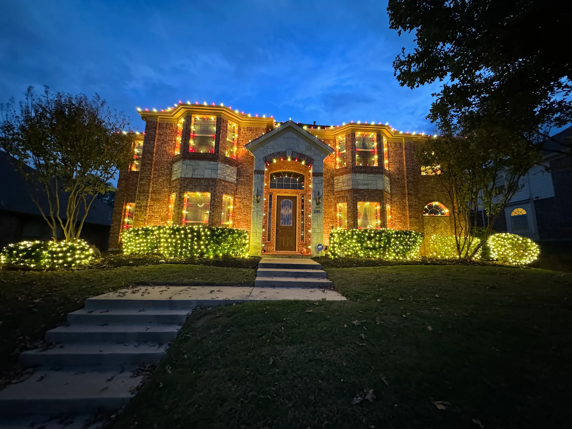 Two-storey brick home photographed at dusk, with the roofline and every window outlined in red and warm white C9 bulbs and net lights across the front hedges.