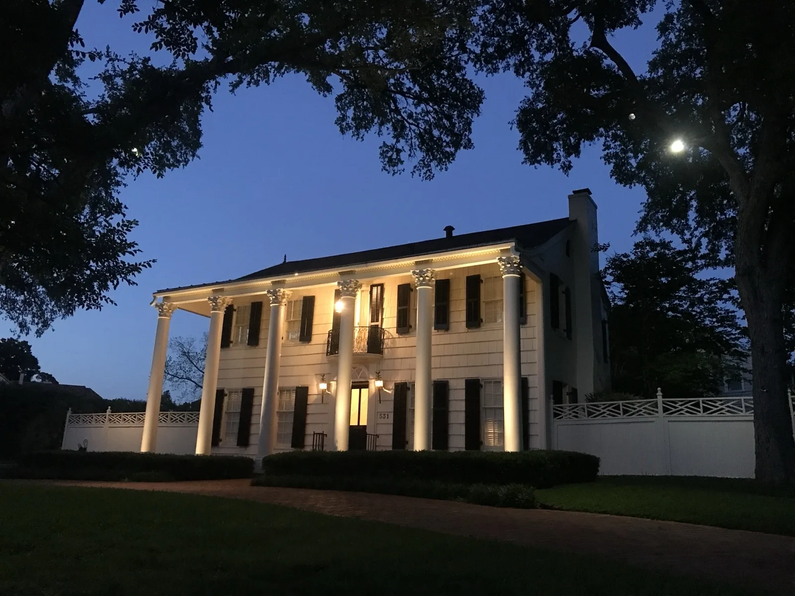 Two-story white house with tall columns, lit at dusk under large trees in Terrell Hills