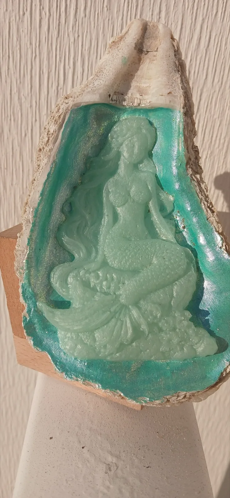 Oyster shell with a pale mermaid figure on a turquoise center, hanging against a light wall