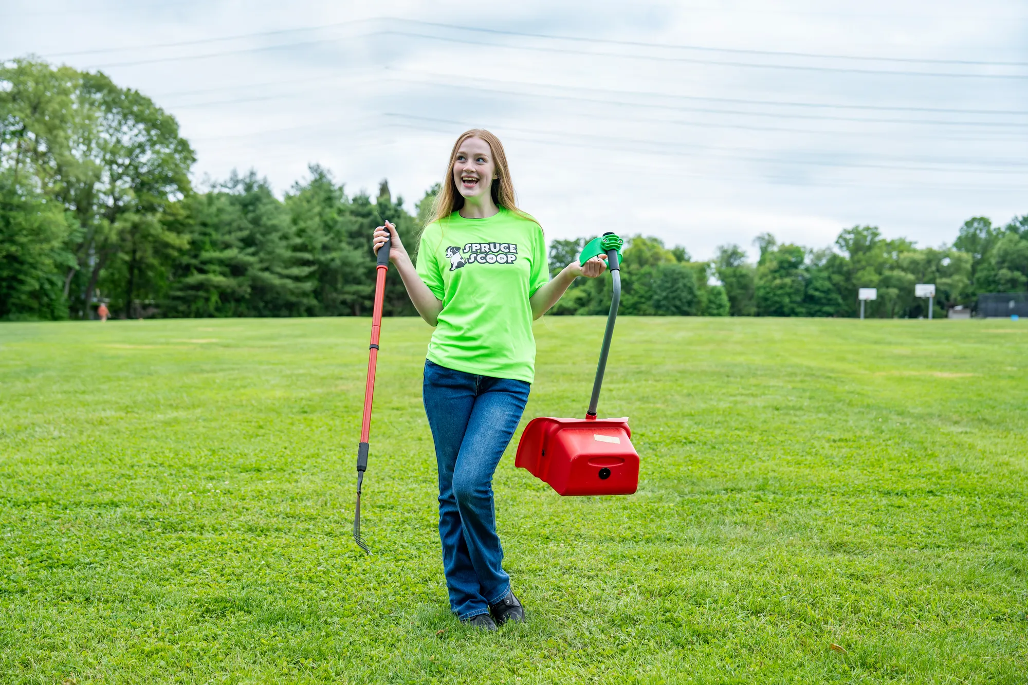 A smiling Spruce Scoop team member holding up a rake and a red scooper pan on a grassy field