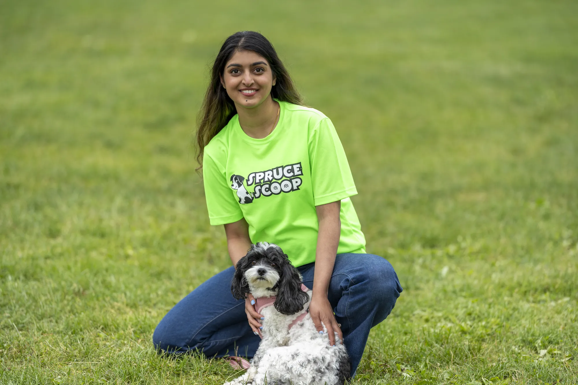 A smiling Spruce Scoop team member kneeling on the grass with a small black-and-white dog