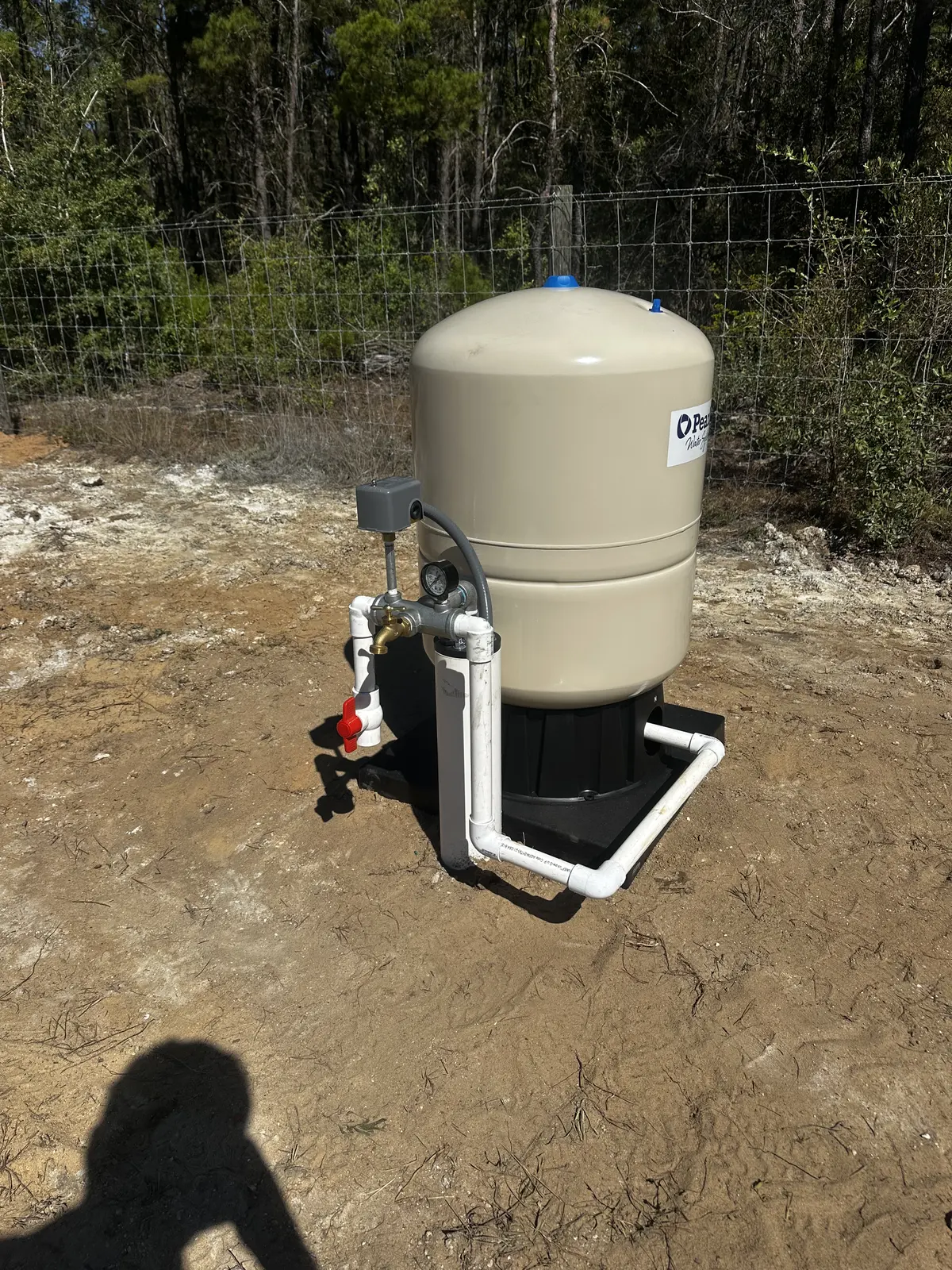Beige water pressure tank with valves and white PVC piping installed on a sandy site
