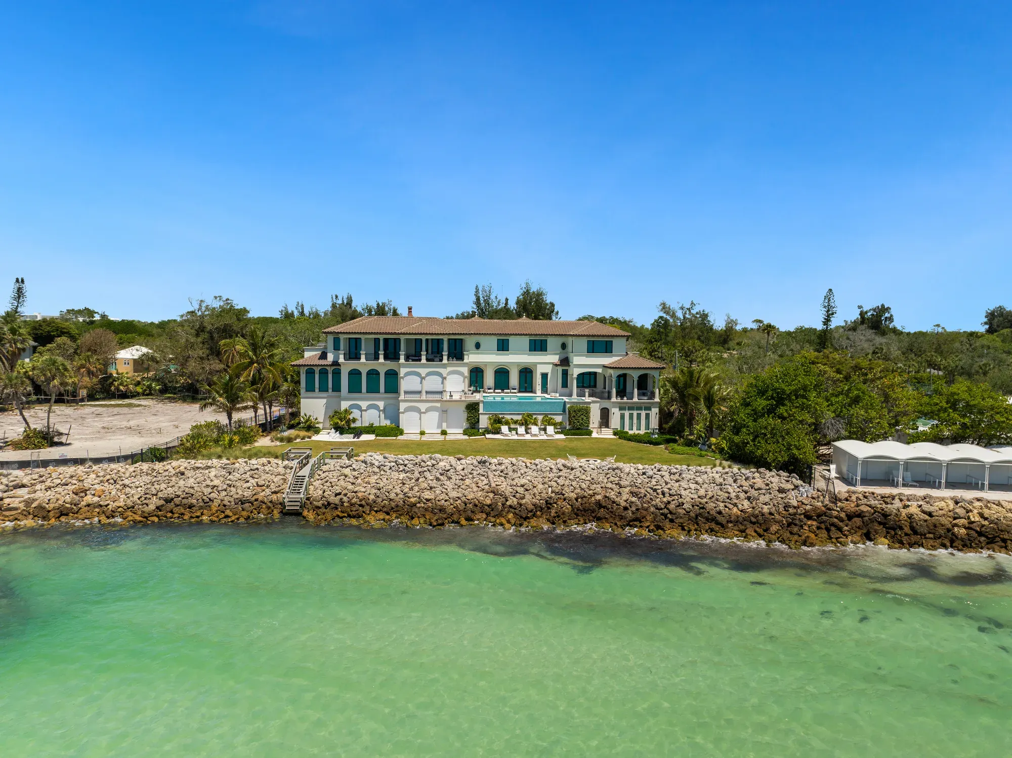 A stunning waterfront mansion features a blend of greenery and rocky shoreline. The spacious property boasts large windows, a pool area, and palm trees, situated against a clear blue sky.