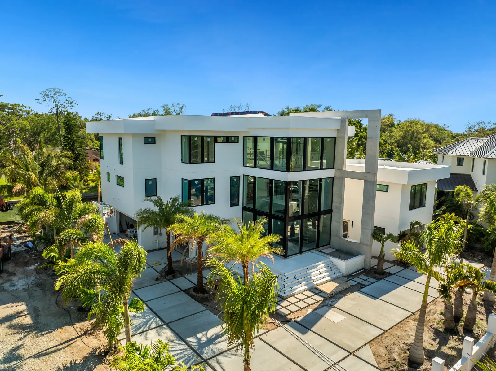 A contemporary multi-story house featuring large windows and a sleek design, surrounded by palm trees. The property includes a spacious driveway and is set against a clear blue sky.