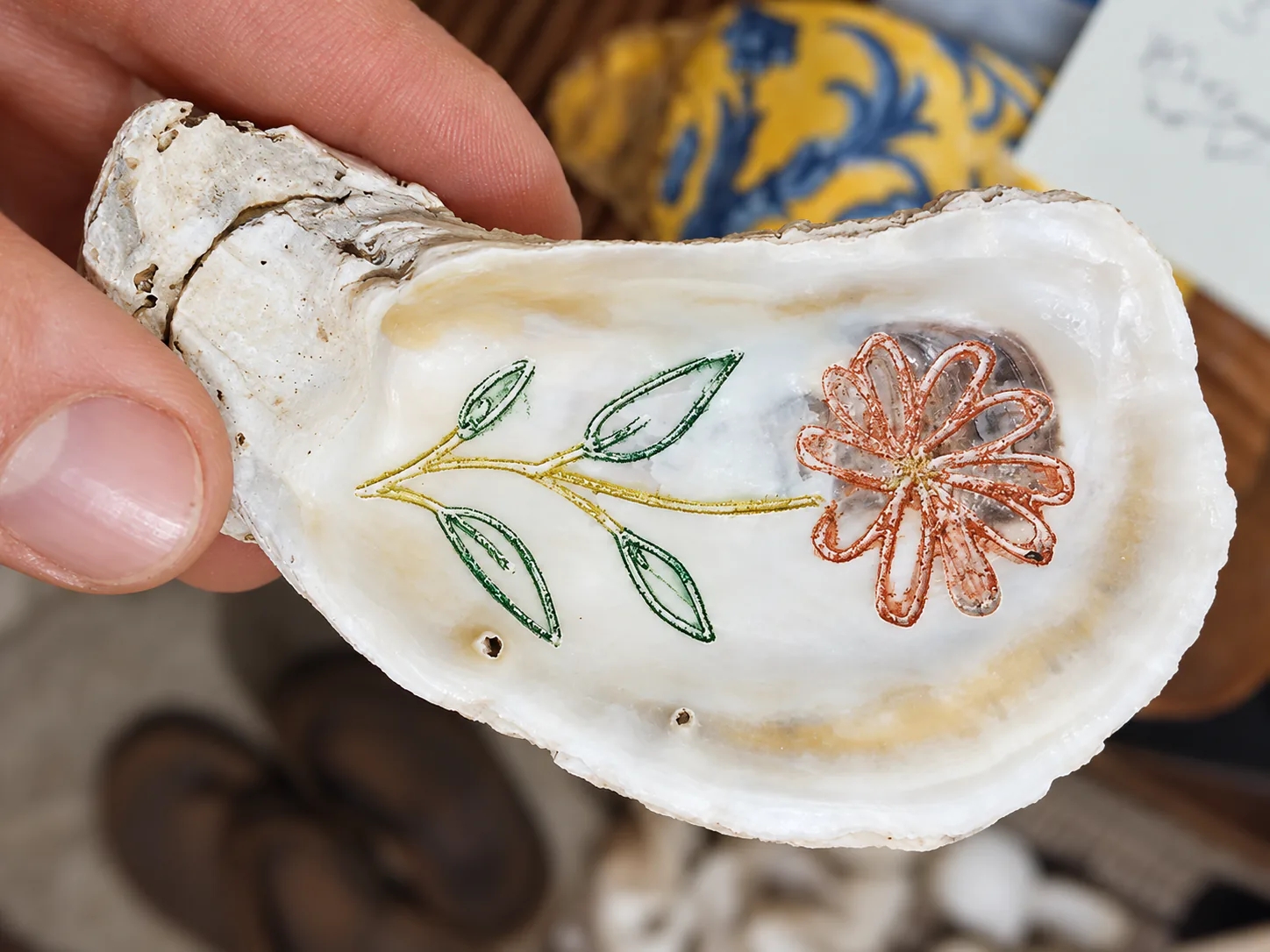 Hand holding an oyster shell engraved with a flower and leaves in red, yellow and green