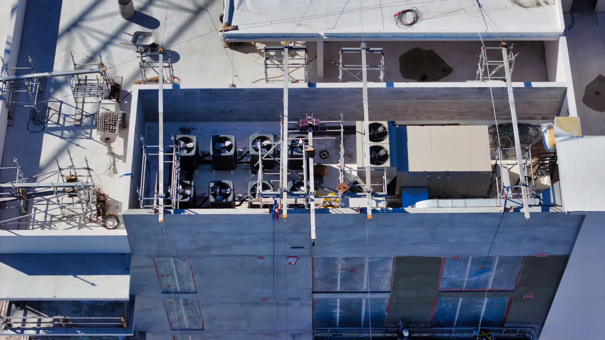 An aerial view of a construction site rooftop displaying scaffolding, cooling units, and various construction materials. The site features several air conditioning units and equipment positioned on the flat roof amidst scaffolding for ongoing construction work.