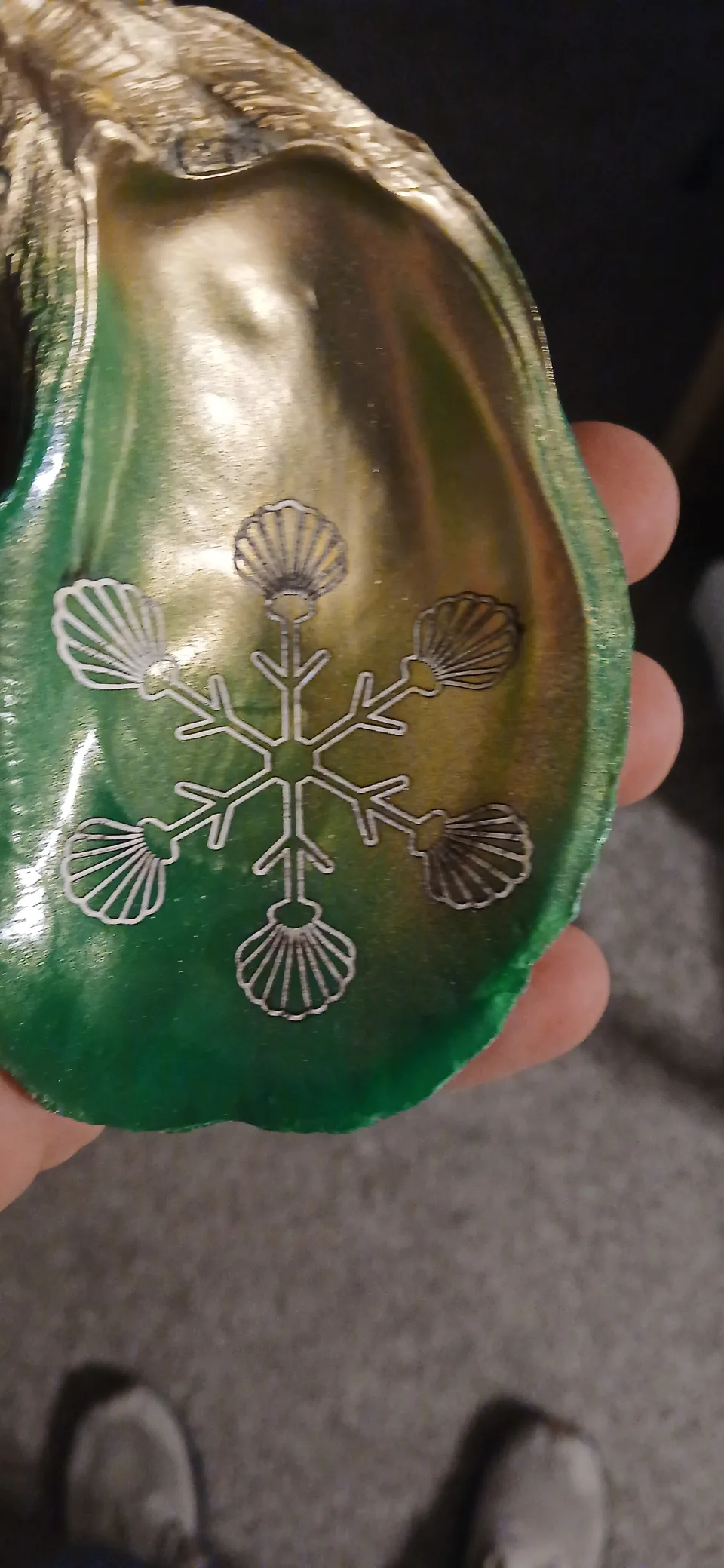 Hand holding a green and gold oyster shell with a snowflake design made from six scallop shapes