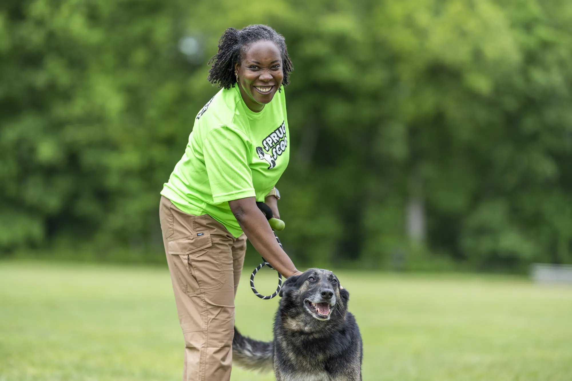A smiling Spruce Scoop team member with a dark dog on a leash on a grassy field