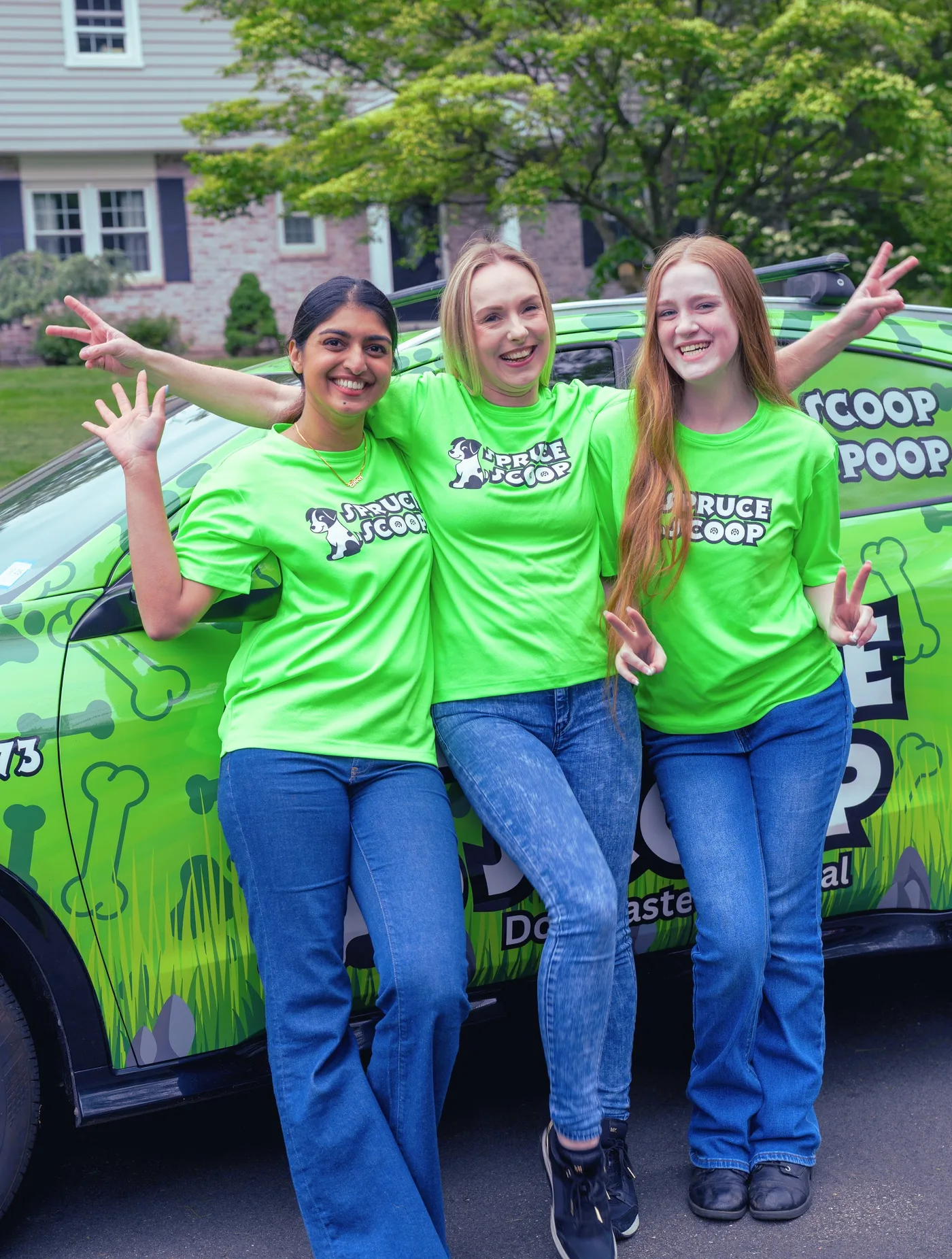 Three smiling Spruce Scoop team members waving and throwing peace signs beside a wrapped Spruce Scoop car