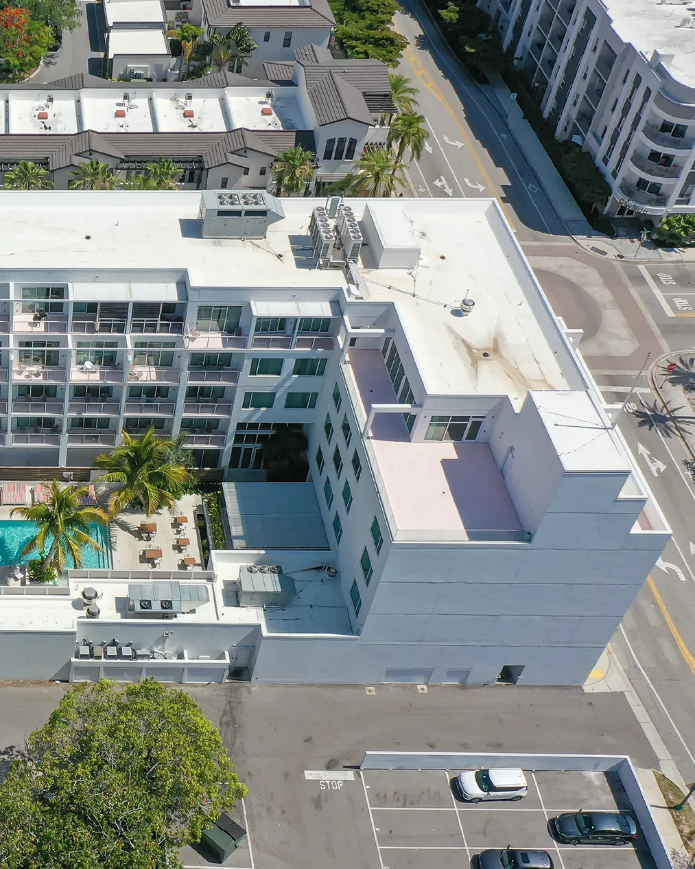 The image depicts an aerial view of a multi-story hotel complex featuring a swimming pool surrounded by lounge chairs. Adjacent buildings and a parking lot are visible, emphasizing the urban setting.