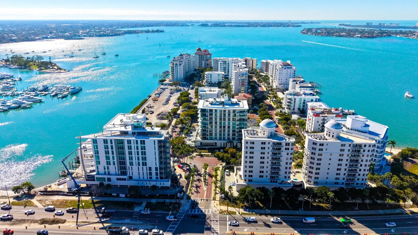 An aerial view of a coastal marina with numerous boats docked, alongside modern residential buildings. The turquoise waters reflect sunlight, creating a scenic backdrop for the developed shoreline.