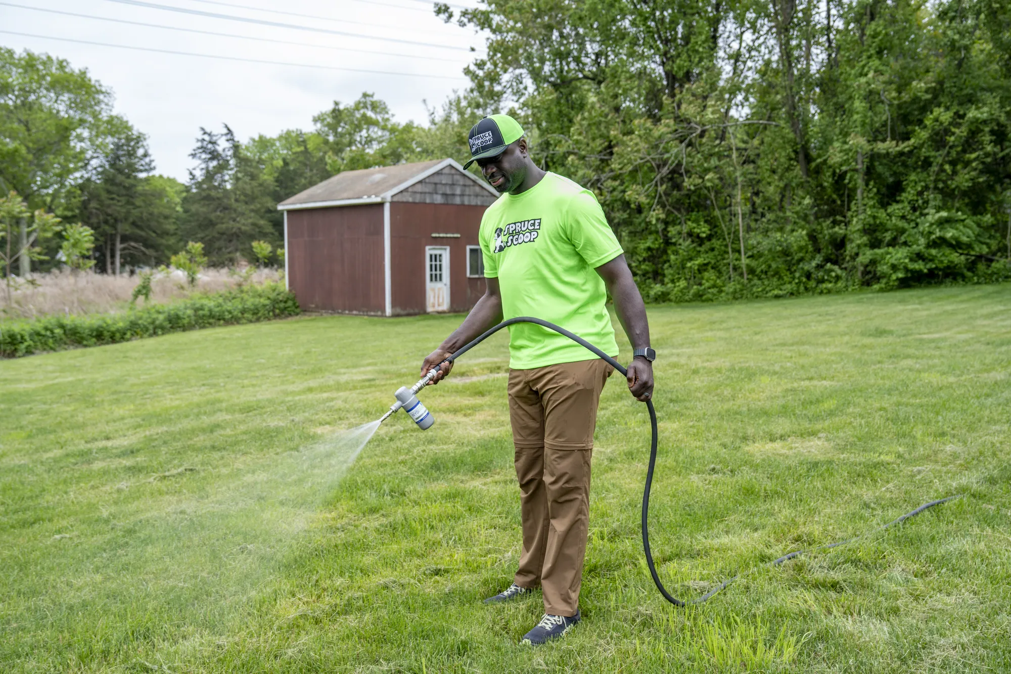 A Spruce Scoop team member in a cap spraying a lawn with a hose-end sprayer near a red shed