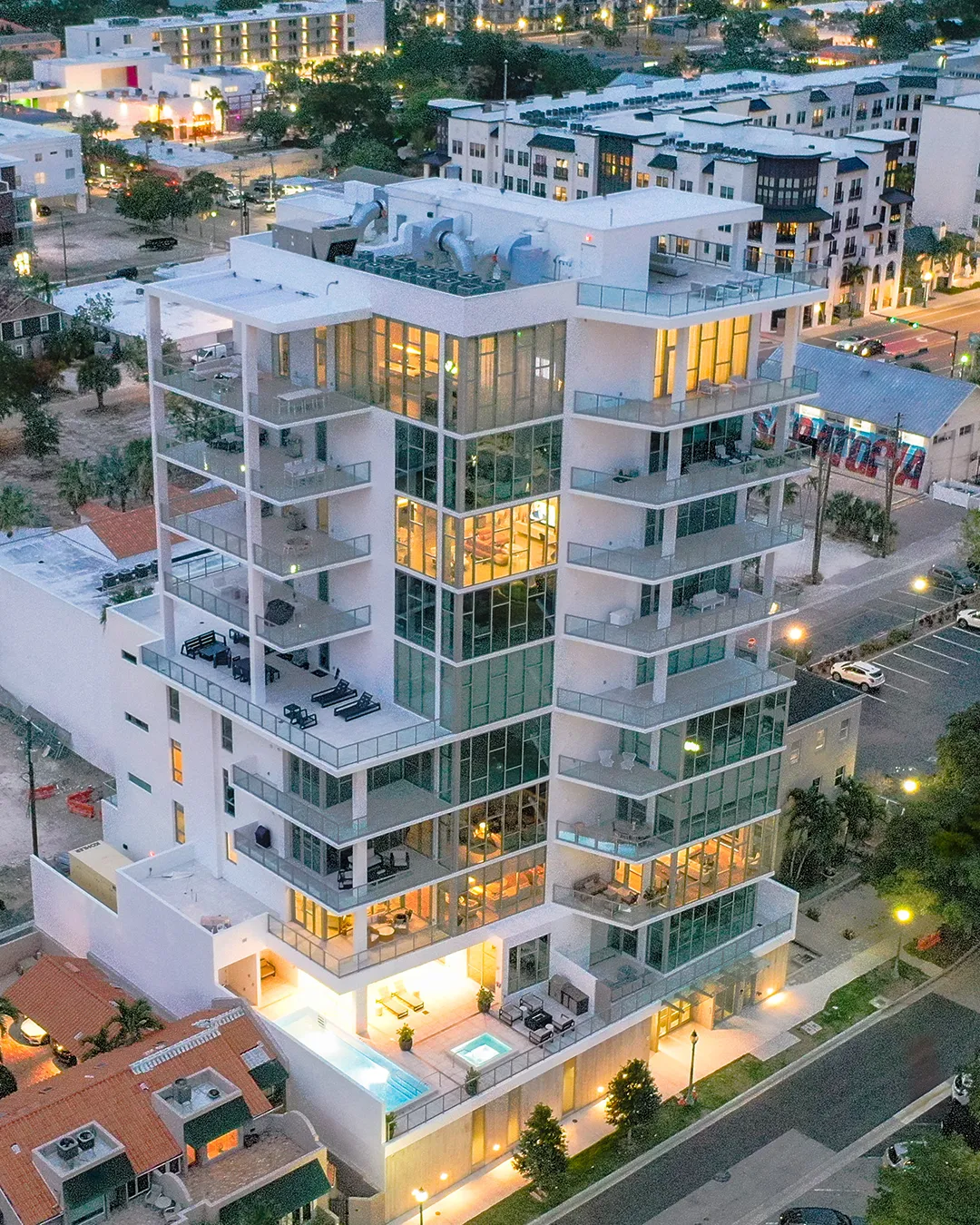 An aerial view of a sleek, modern high-rise building with large glass windows and multiple balconies, illuminated in the evening. The structure features a rooftop and amenities, set against a backdrop of city lights and streets.