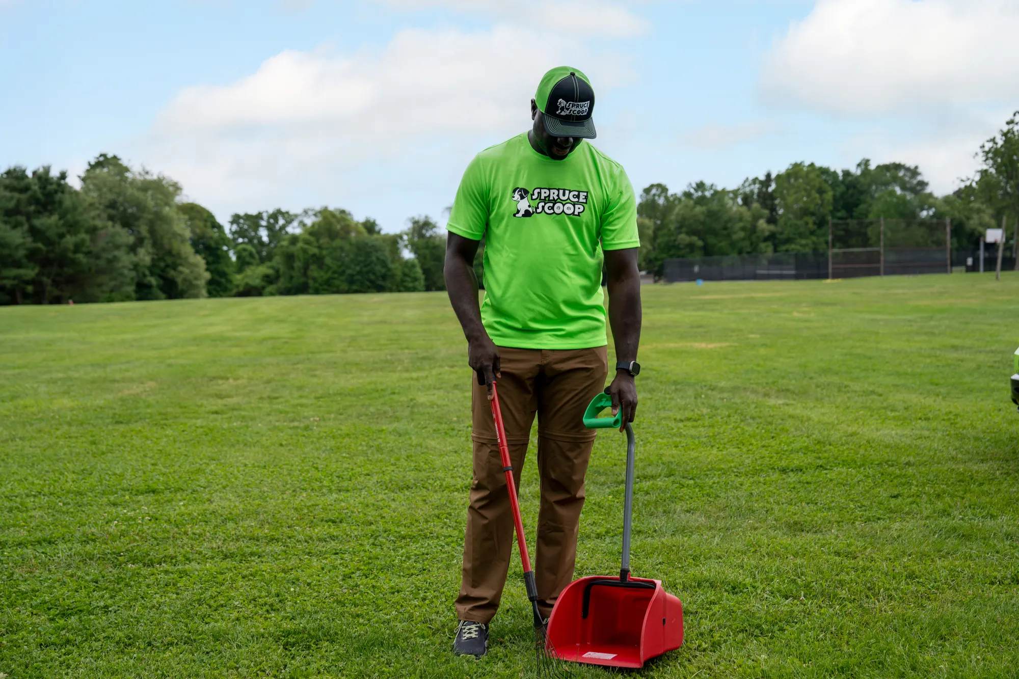 A Spruce Scoop team member in a cap standing on a grassy field with a rake and a red scooper pan