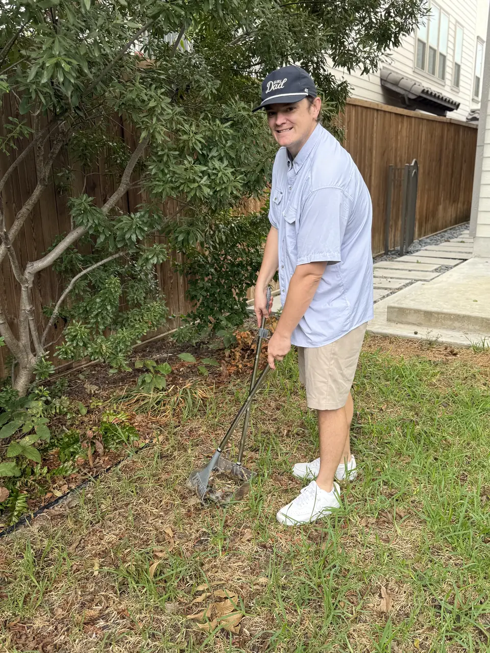 B.P. Foley scooping pet waste with a rake and pan beside a shrub and a wooden fence
