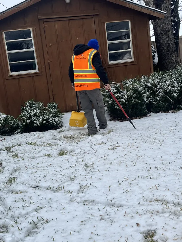 Worker in reflective safety vest shoveling snow in front of a wooden shed