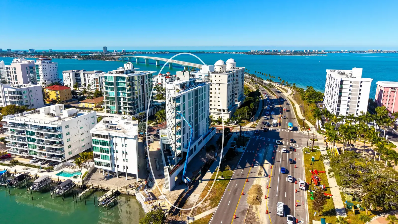 An aerial view of a coastal city featuring modern high-rise buildings along a waterway, with construction work visible on one of the structures. The scene includes vehicles on a roadway, palm trees, and a clear blue sky, highlighting a vibrant urban environment.