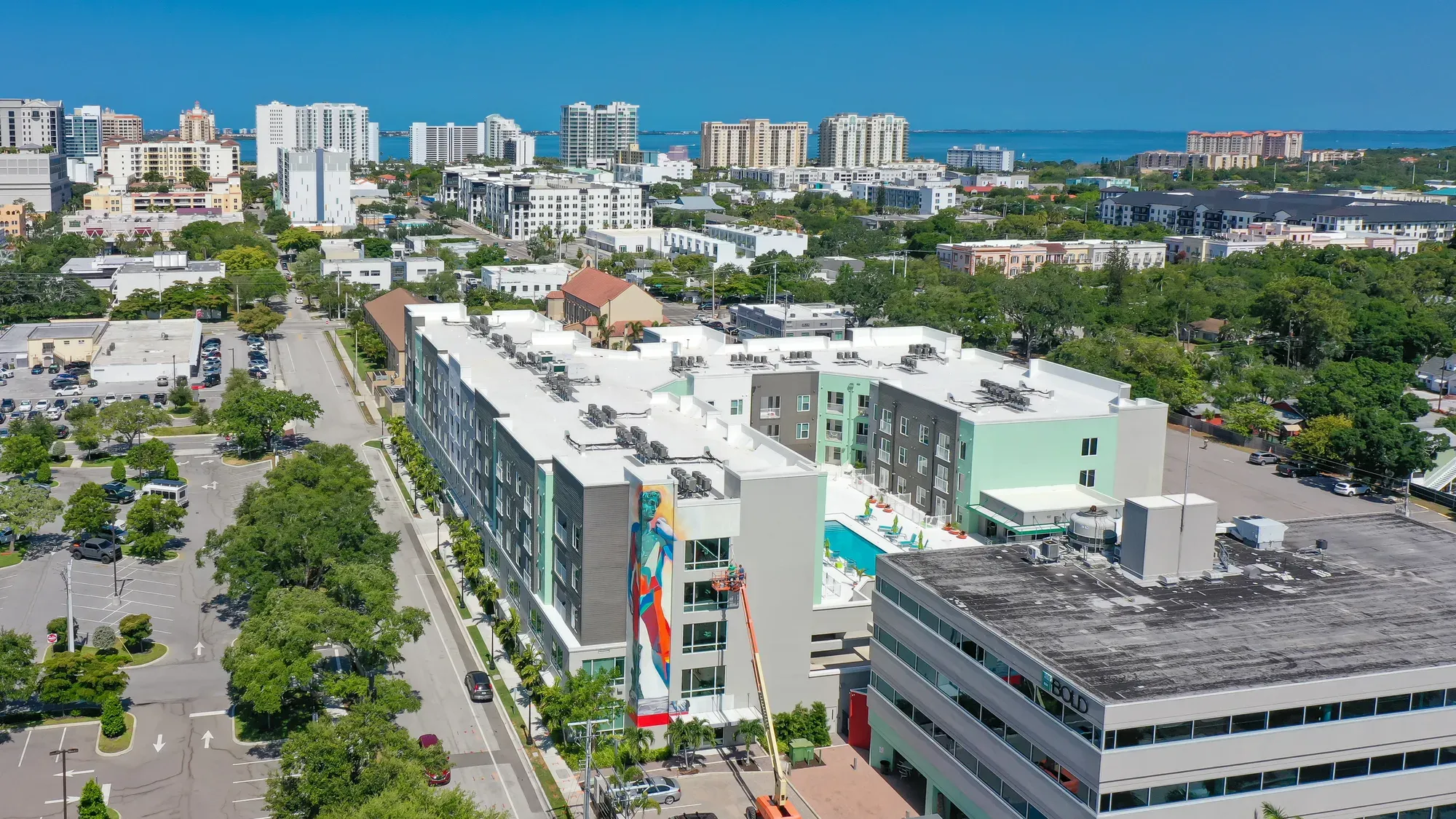 The image shows an aerial view of a modern urban area featuring a recently constructed building with a pool, vibrant mural, and surrounding greenery. In the background, a skyline of high-rise buildings and distant water can be seen, indicating a coastal city atmosphere.