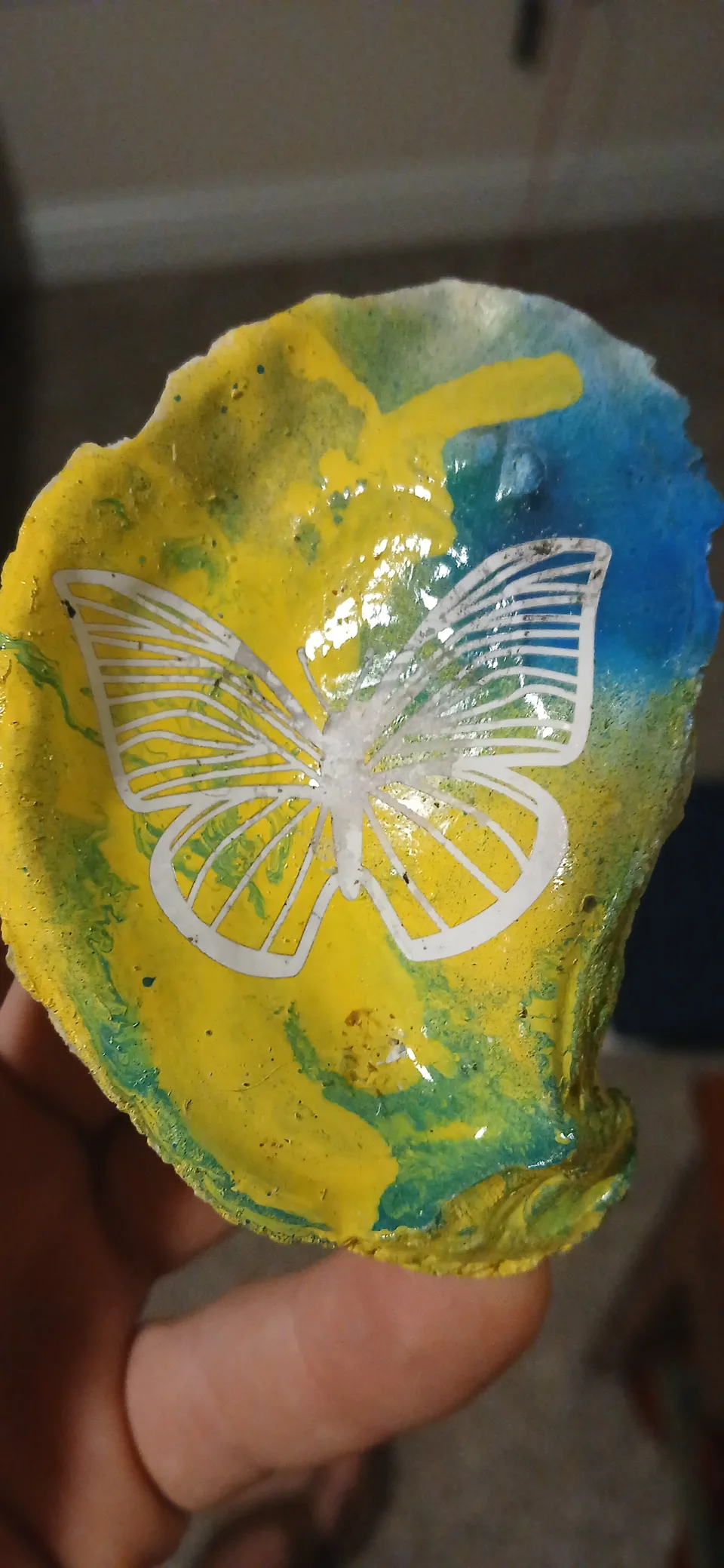 Hand holding a yellow, green and blue oyster shell engraved with a white butterfly