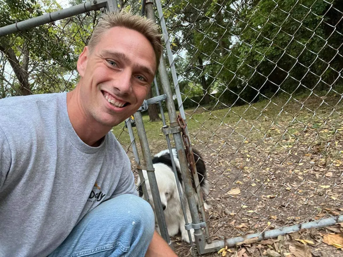 Smiling man taking a selfie near a fence with two dogs peeking out