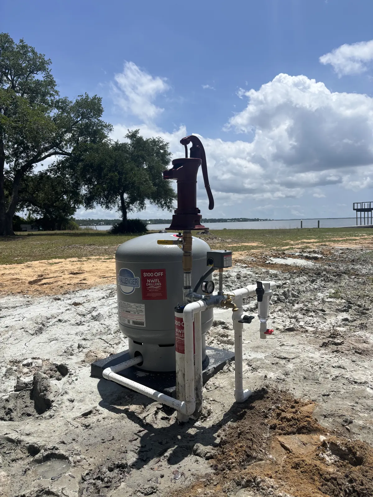 Water pump with a red handle and white PVC piping installed near the waterfront under a blue sky