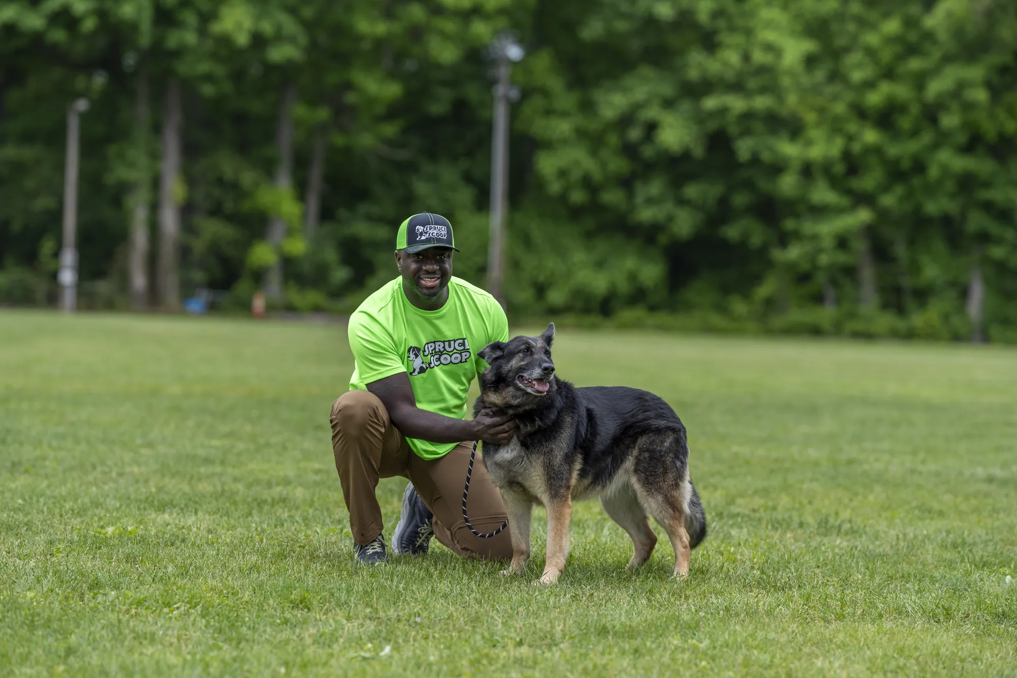 A Spruce Scoop team member in a cap crouching beside a large dark dog on a grassy field
