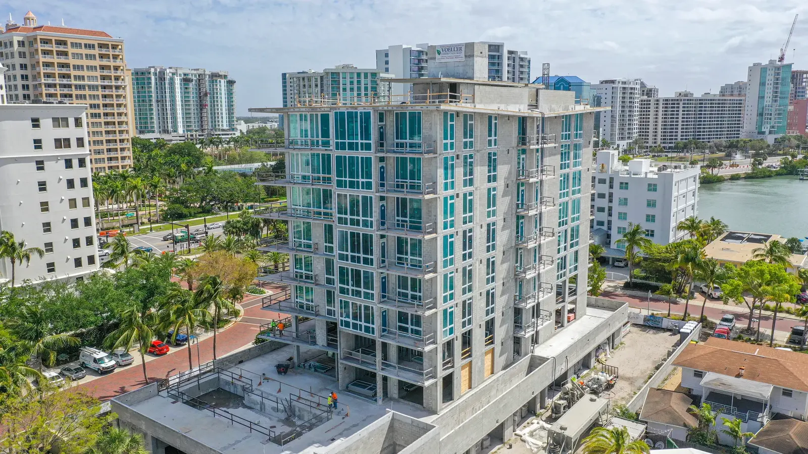 An aerial view of a modern high-rise building under construction, featuring multiple glass windows and a concrete structure. Surrounding the building are palm trees, roads, and other urban developments, showcasing a vibrant city environment.