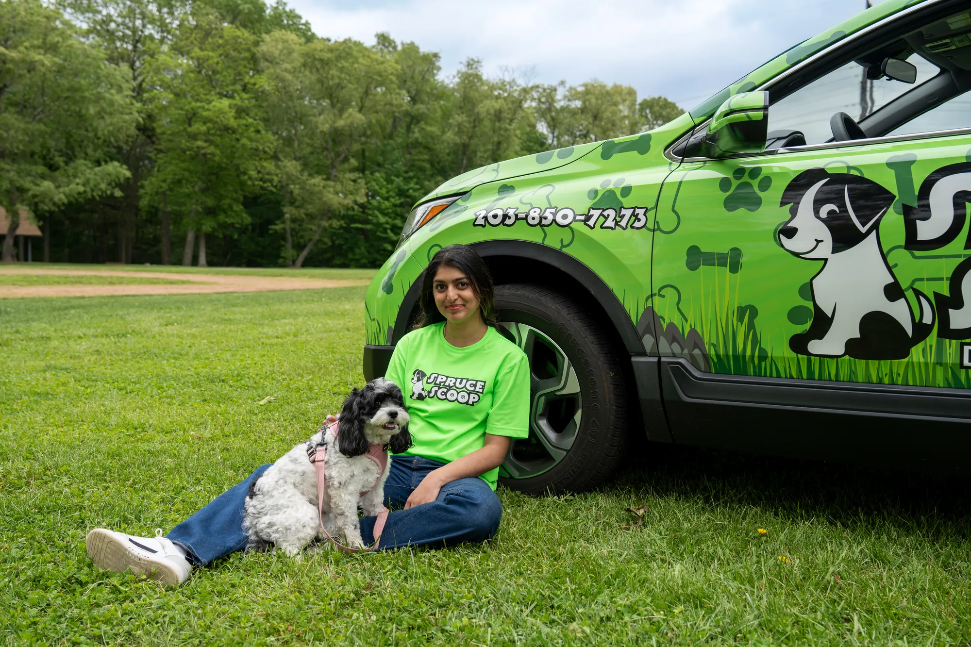 A Spruce Scoop team member sitting on the grass against a wrapped Spruce Scoop car with a small fluffy dog