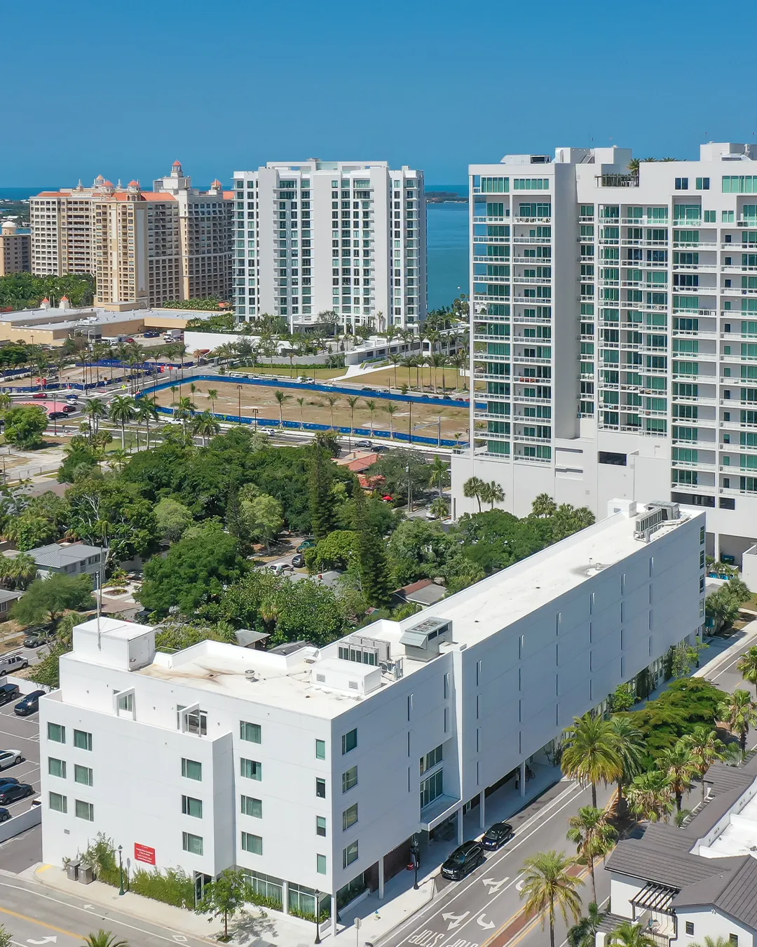 An aerial view of a modern urban area featuring a white multi-story building in the foreground, with taller residential buildings and lush greenery in the background, alongside a glimpse of the ocean in the distance under a clear blue sky.