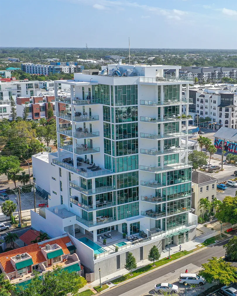 A contemporary high-rise building featuring large glass windows and balconies, set against a clear blue sky. The structure is surrounded by greenery and residential areas, showcasing an urban environment.