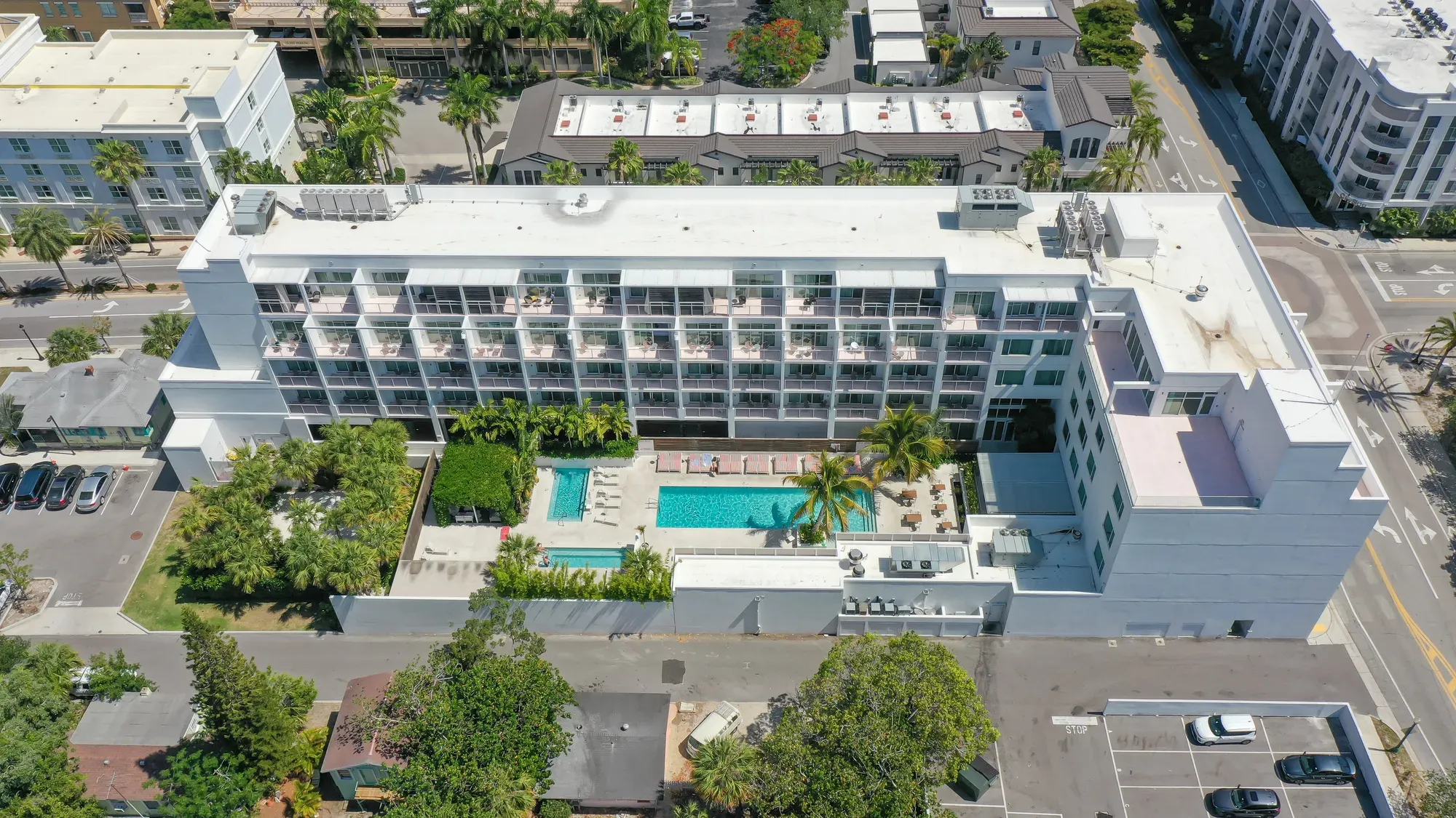 An aerial view of a modern hotel featuring multiple floors, a swimming pool surrounded by lounge chairs, and lush palm trees. The surrounding area includes other buildings and parked cars.