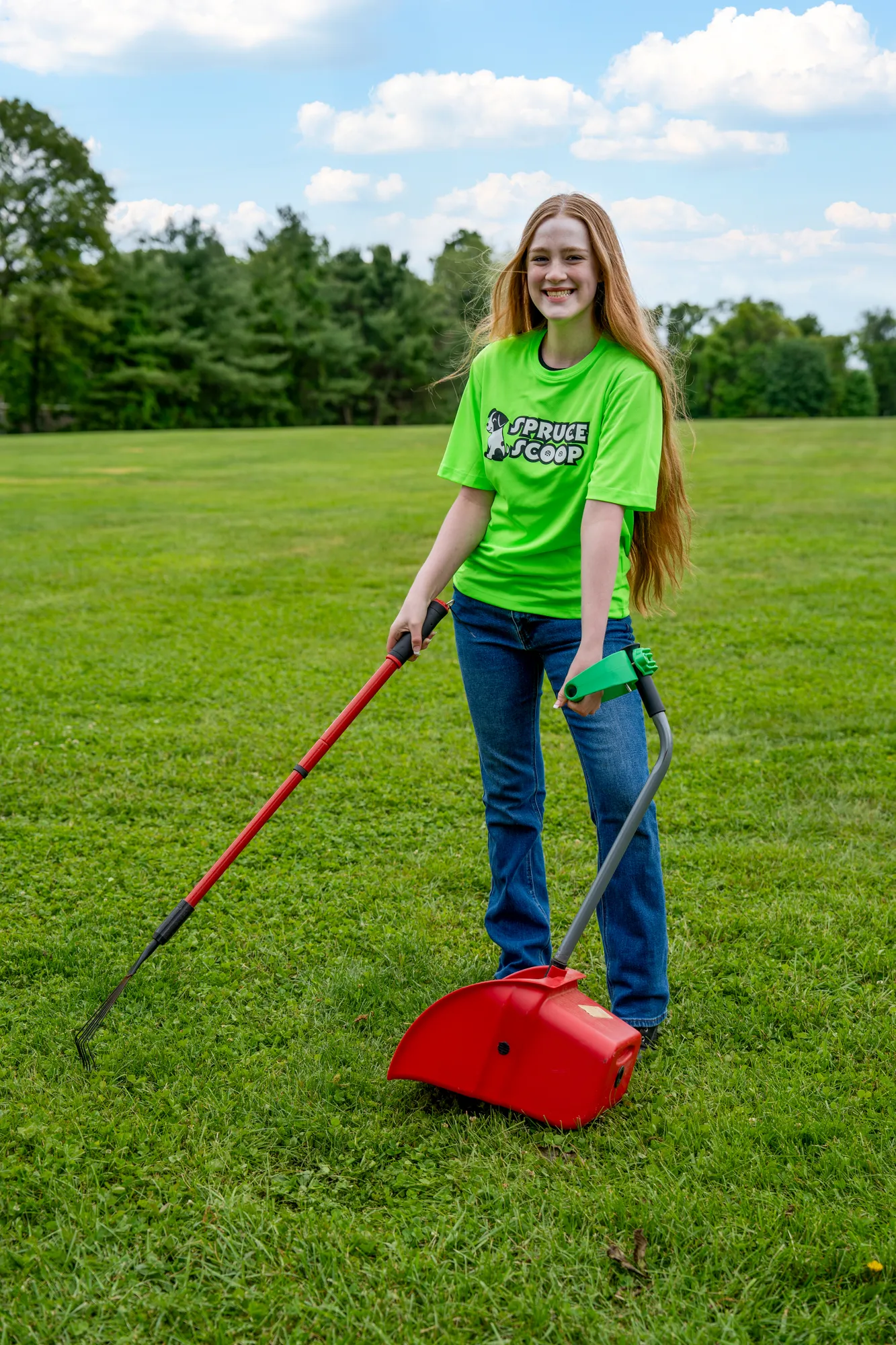 A smiling Spruce Scoop team member using a rake and a red scooper pan on a grassy field
