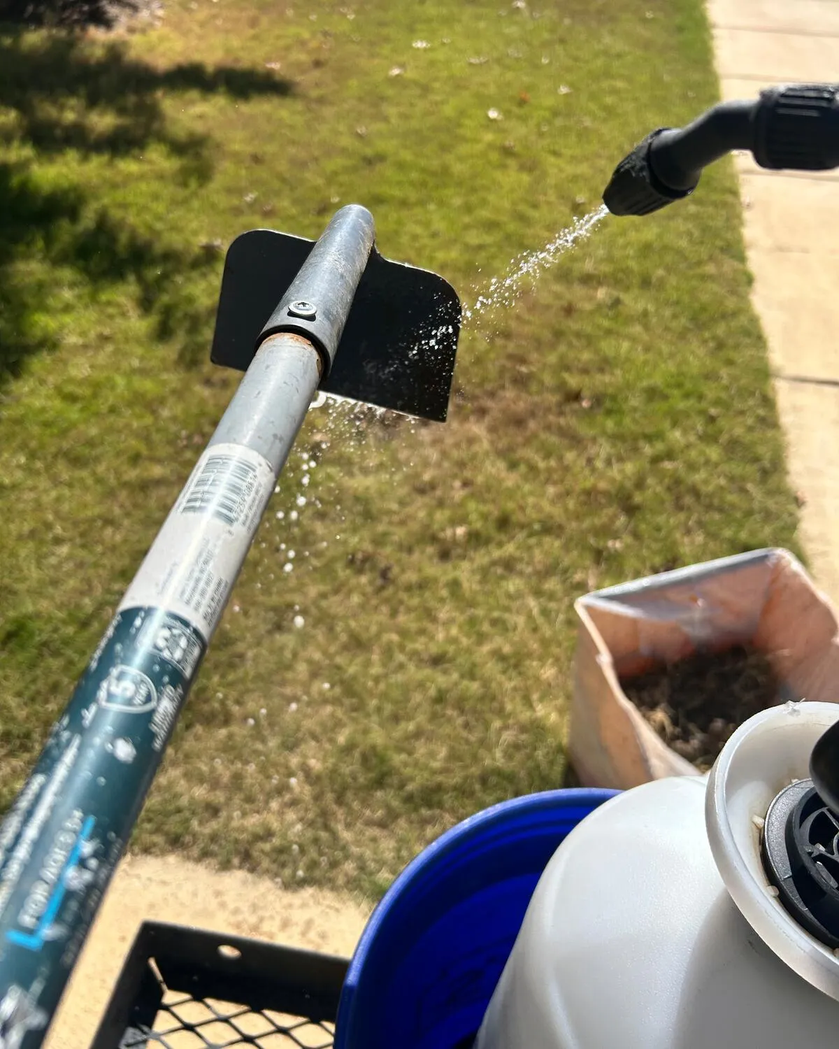 A scoop being rinsed with a sprayer over the grass, above a bucket and a sprayer tank