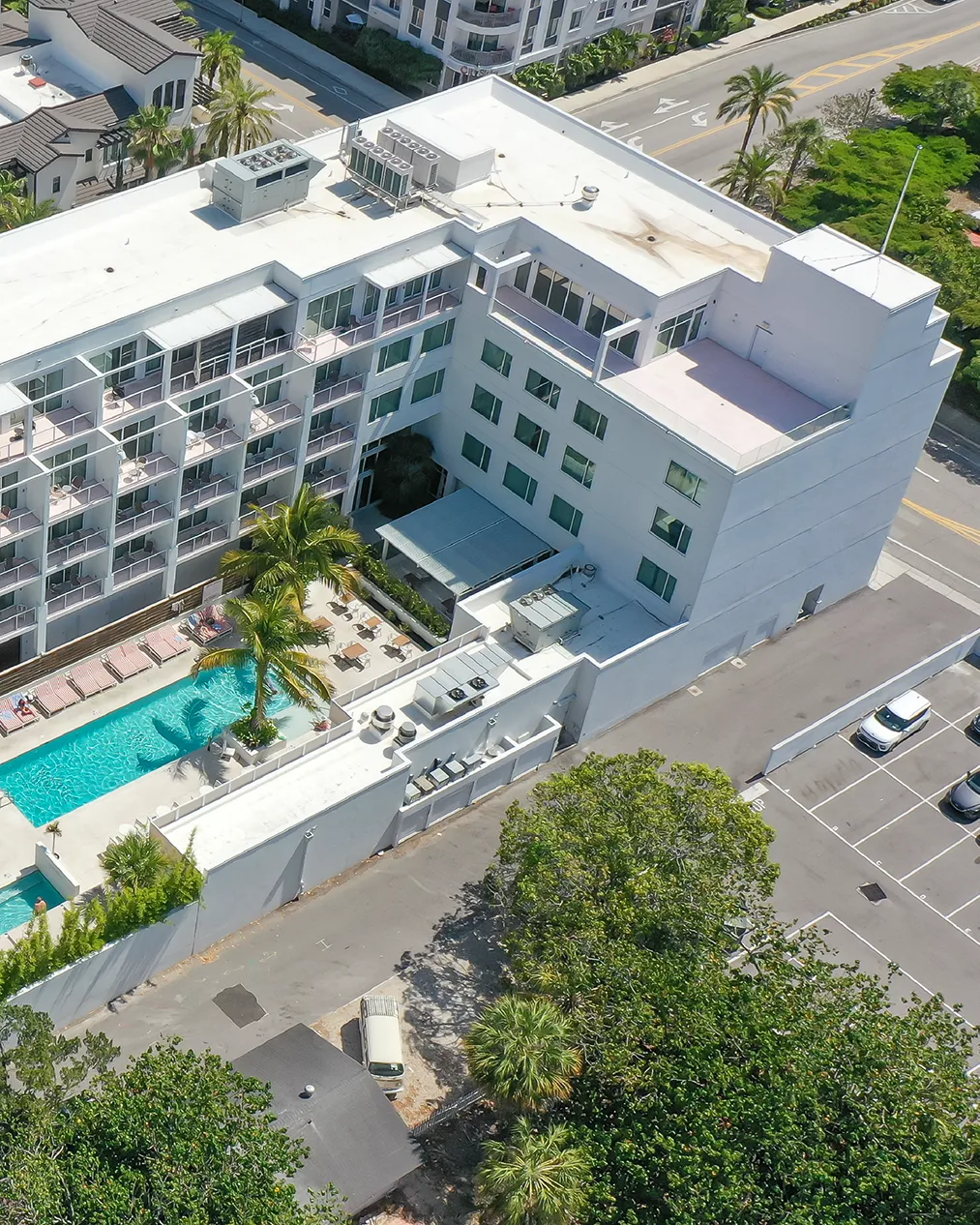 The image depicts an aerial view of a modern hotel featuring a swimming pool surrounded by lounge chairs and palm trees. Adjacent to the hotel are several parking spaces and nearby greenery, along with a roadway in the background.