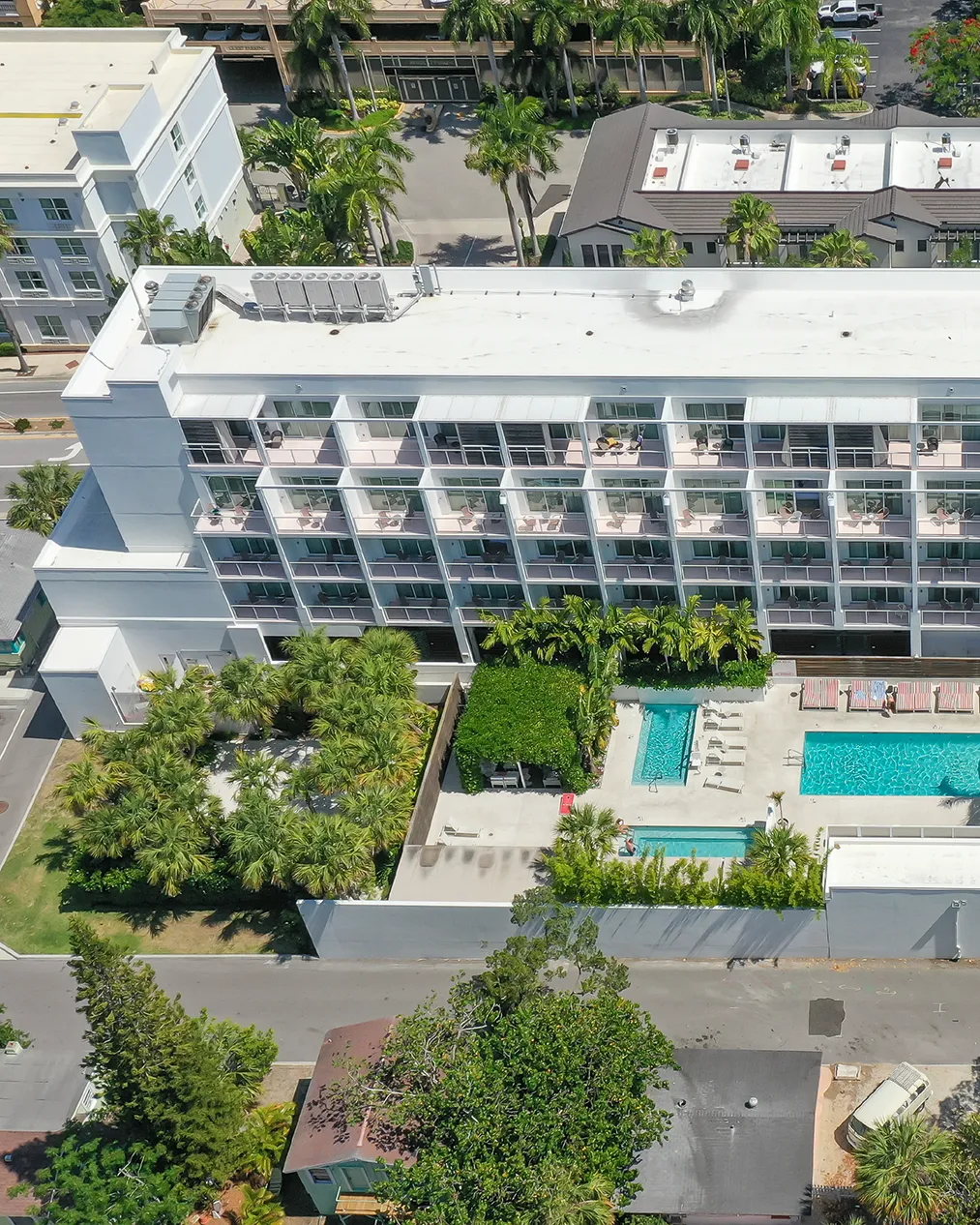 An aerial perspective of a modern hotel featuring multiple floors and balconies, surrounded by lush greenery. The image shows a swimming pool area with lounge chairs and palms, highlighting the tranquil setting.