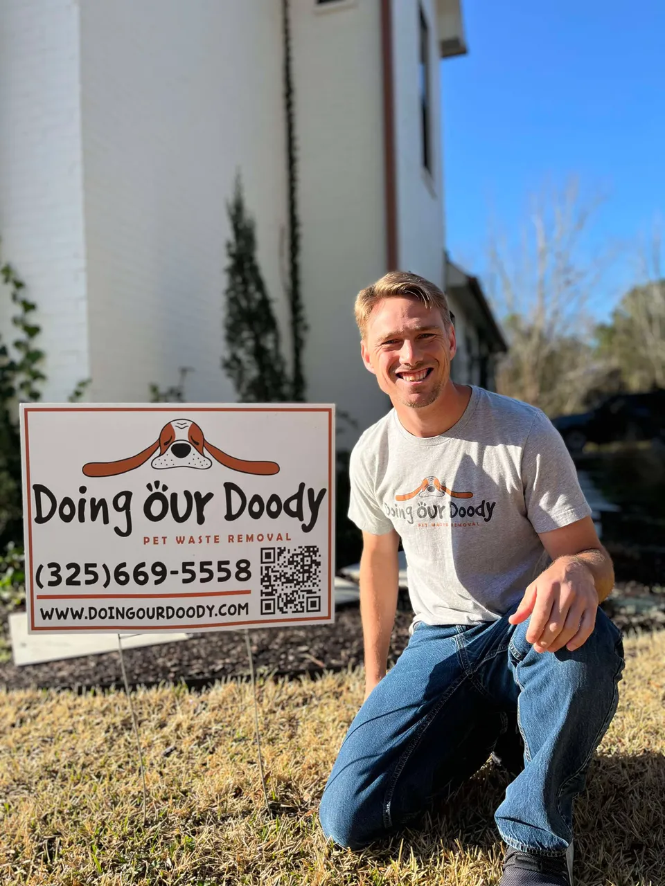 Doing Our Doody owner kneeling beside a yard sign while wearing a branded shirt, with a residential home in the background