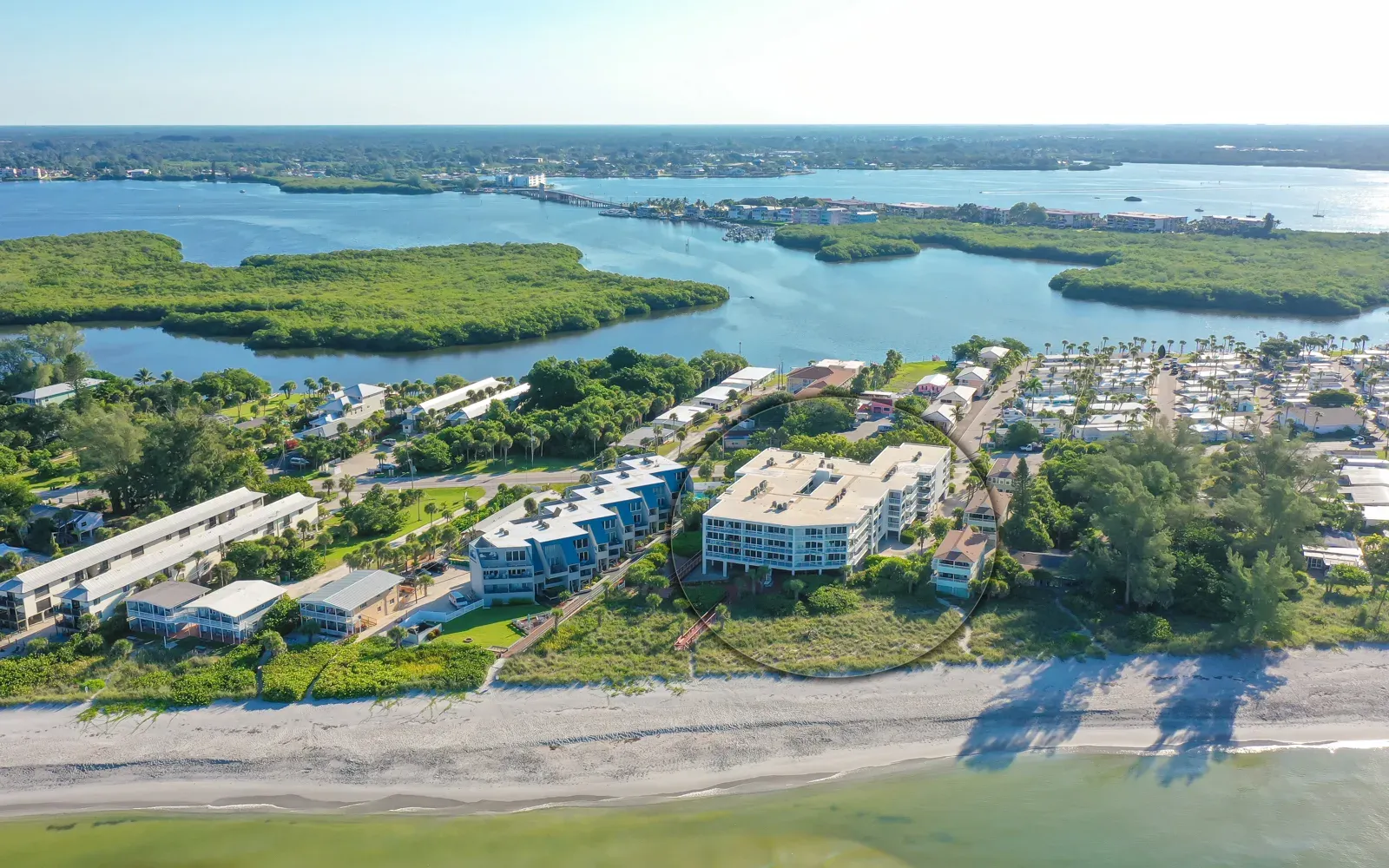 An aerial view of a coastal area featuring residential buildings along the beach, lush greenery, and waterways. The scene captures a blend of waterfront properties and serene natural landscapes.