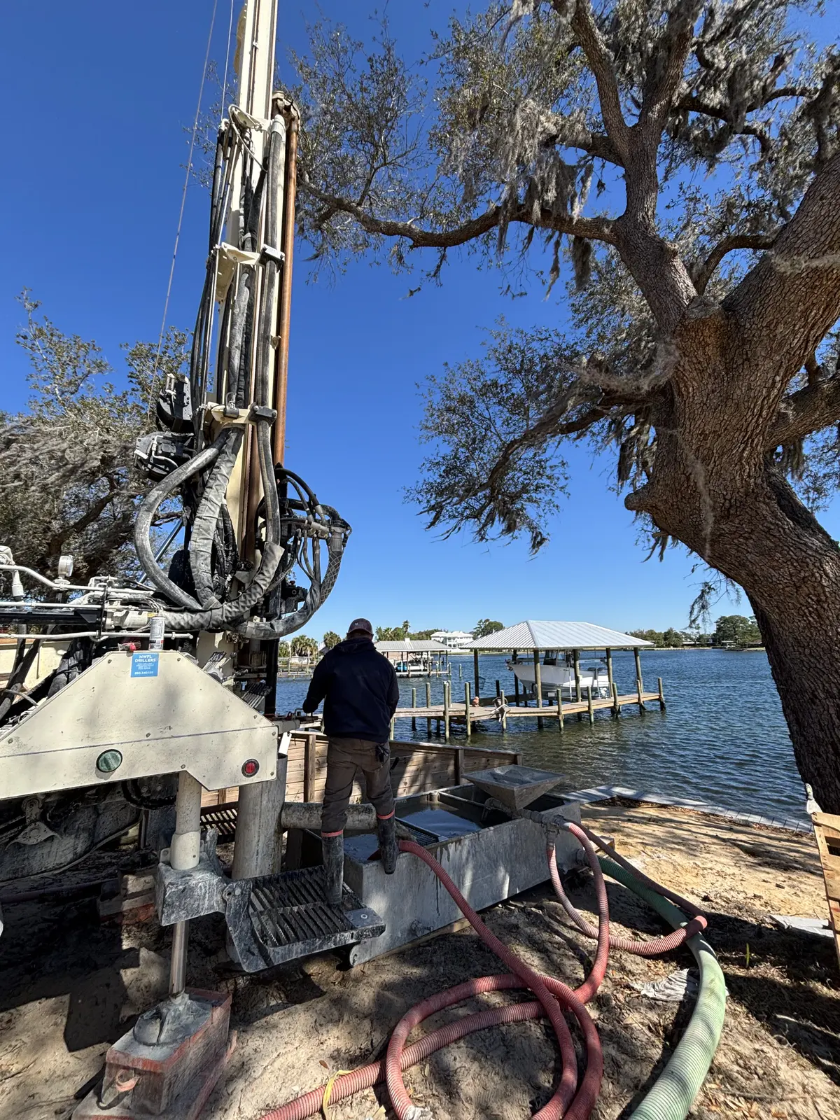Worker operating a water well drilling rig beside the water with hoses running toward the shore